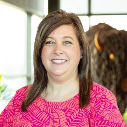 Smiling woman with straight brown hair in a vibrant pink patterned top, posing in front of a buffalo statue indoors.