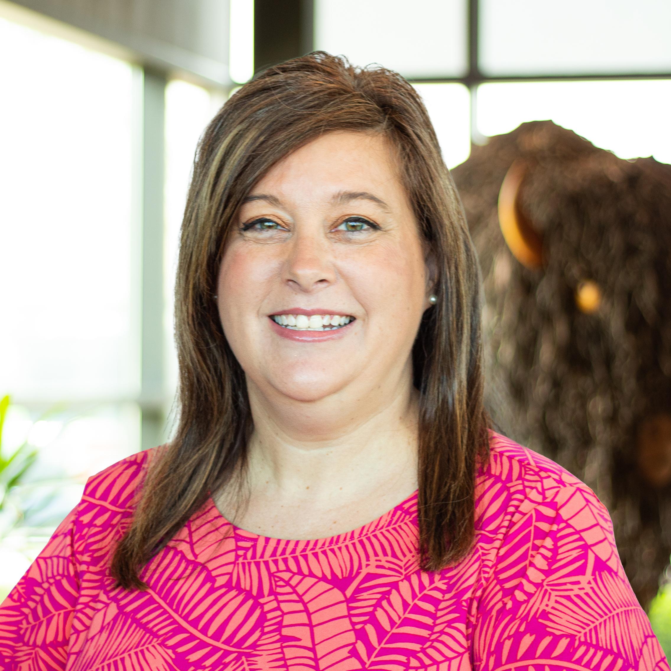 Smiling woman with straight brown hair in a vibrant pink patterned top, posing in front of a buffalo statue indoors.