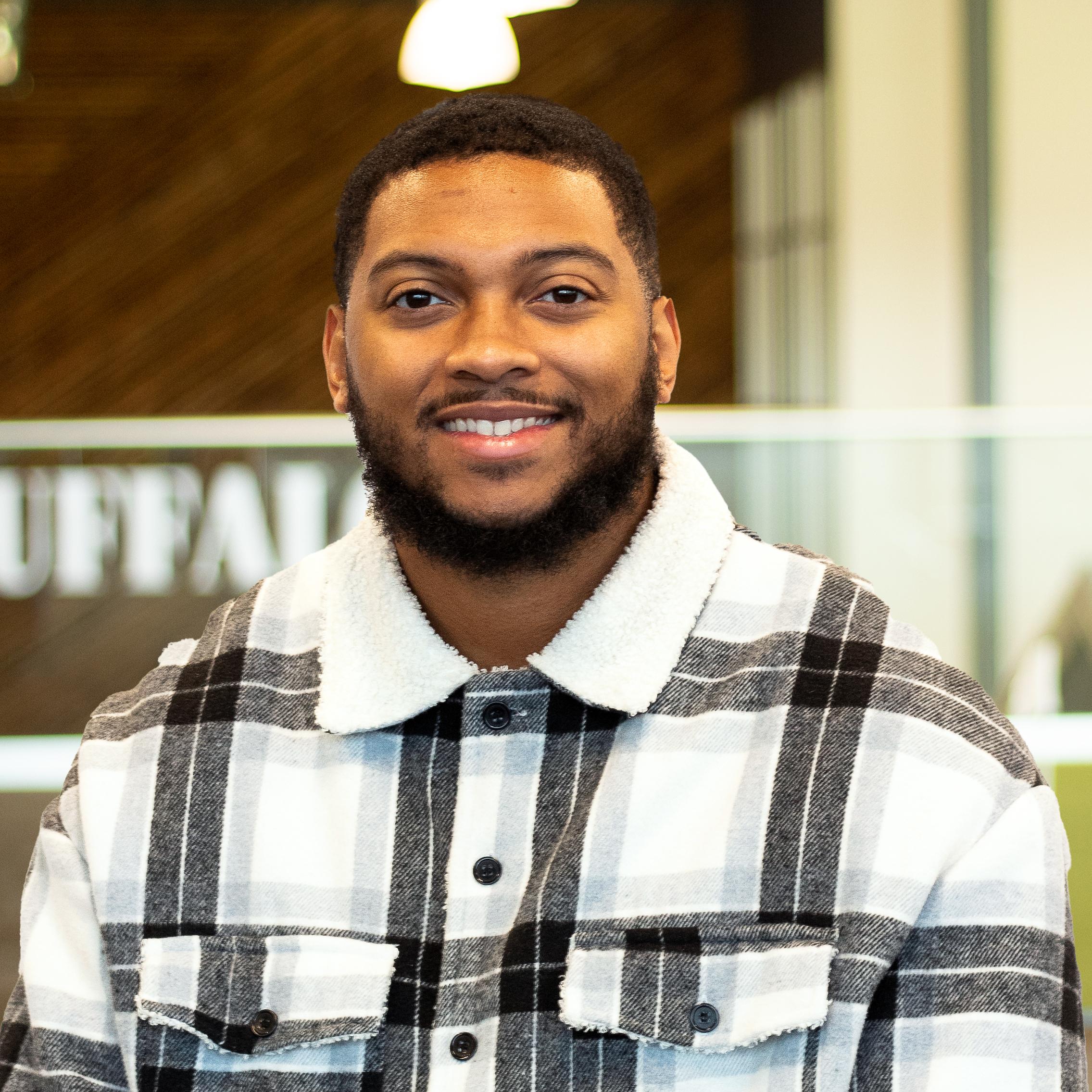 Major Bell smiling, wearing a plaid button down , standing in buffalo's office space.