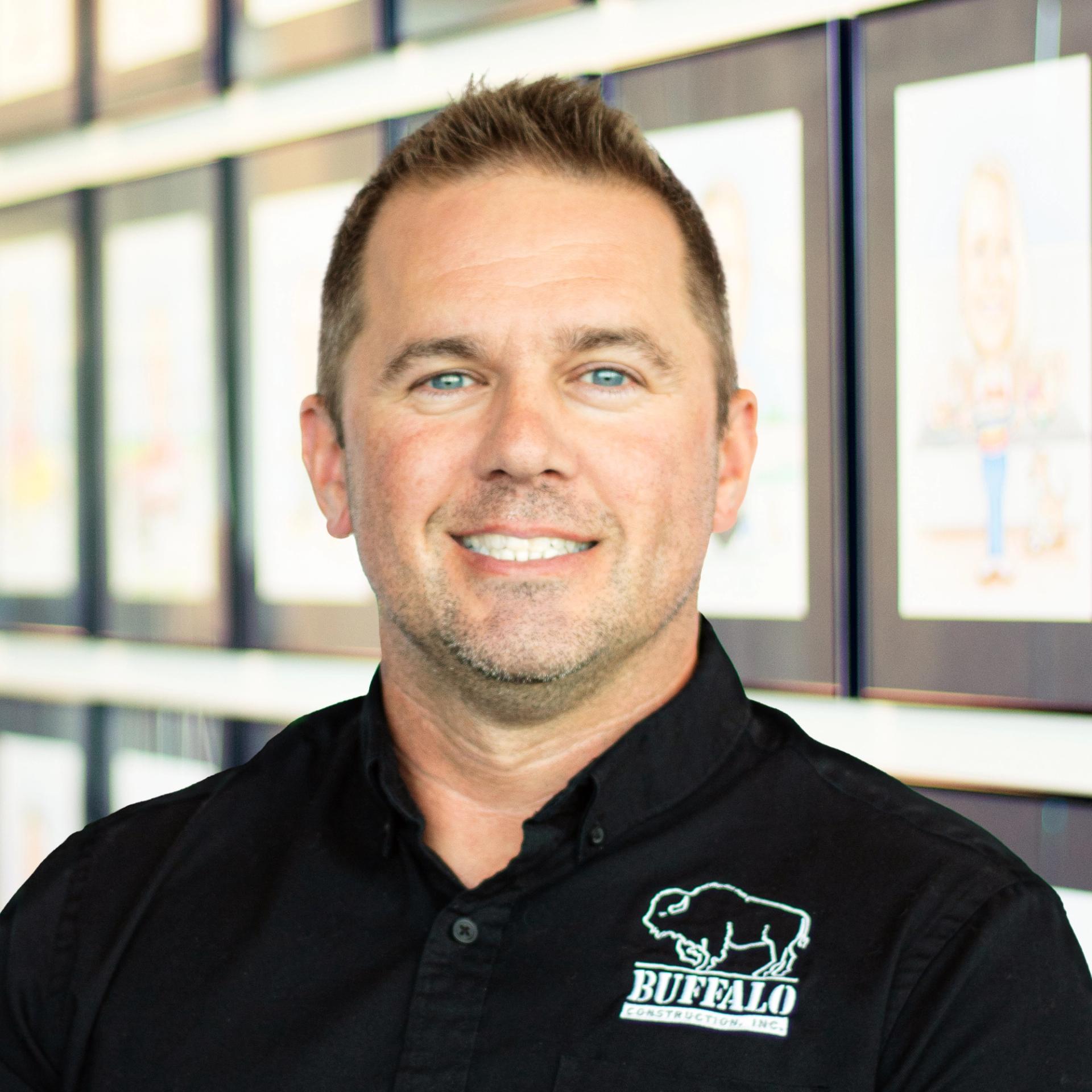 Brian McGee wearing a black buffalo branded polo, smiling and standing in front of a wall displaying framed caricatures