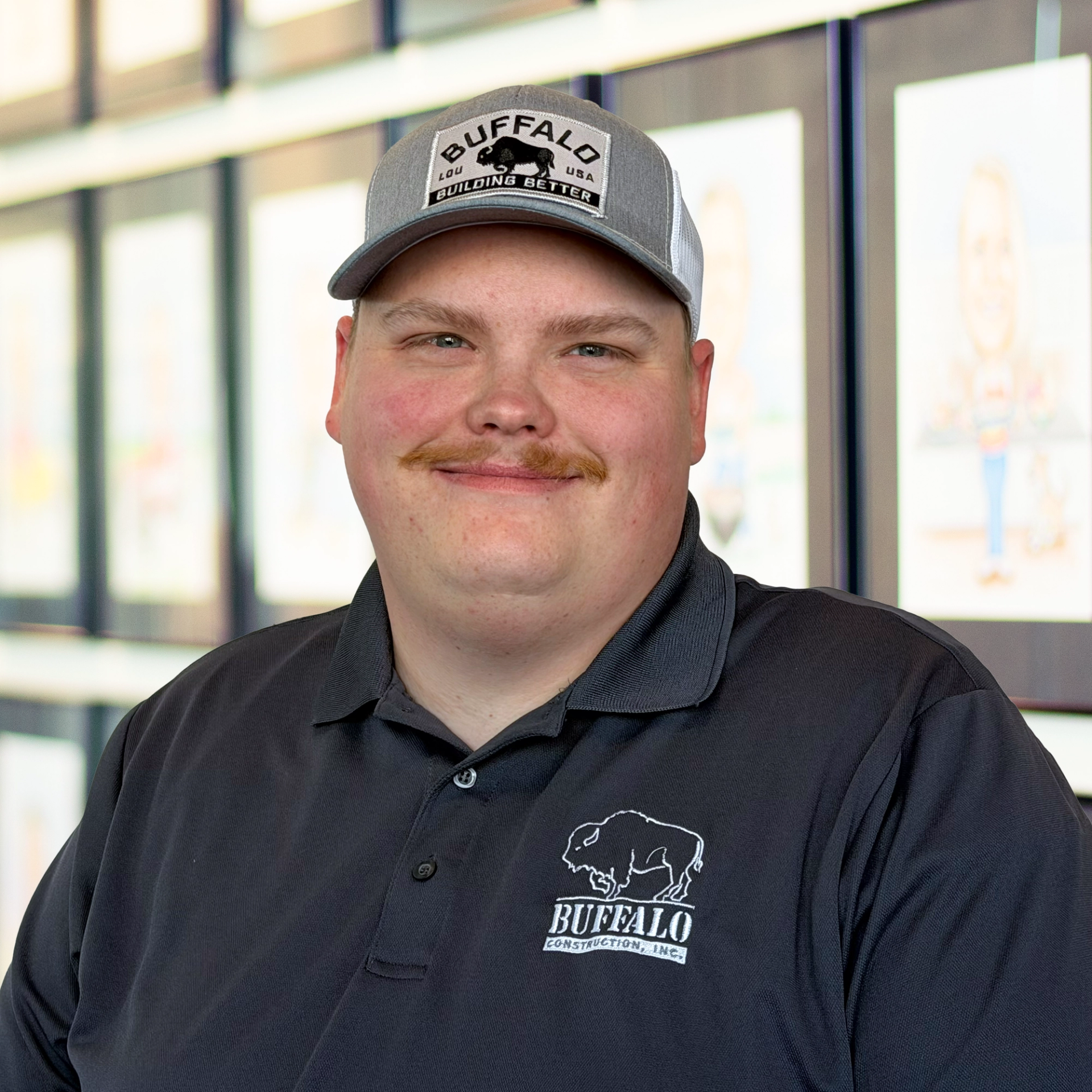 Andrew Weddle, wearing a gray Buffalo hat and black polo, standing in front of a wall displaying framed caricatures.