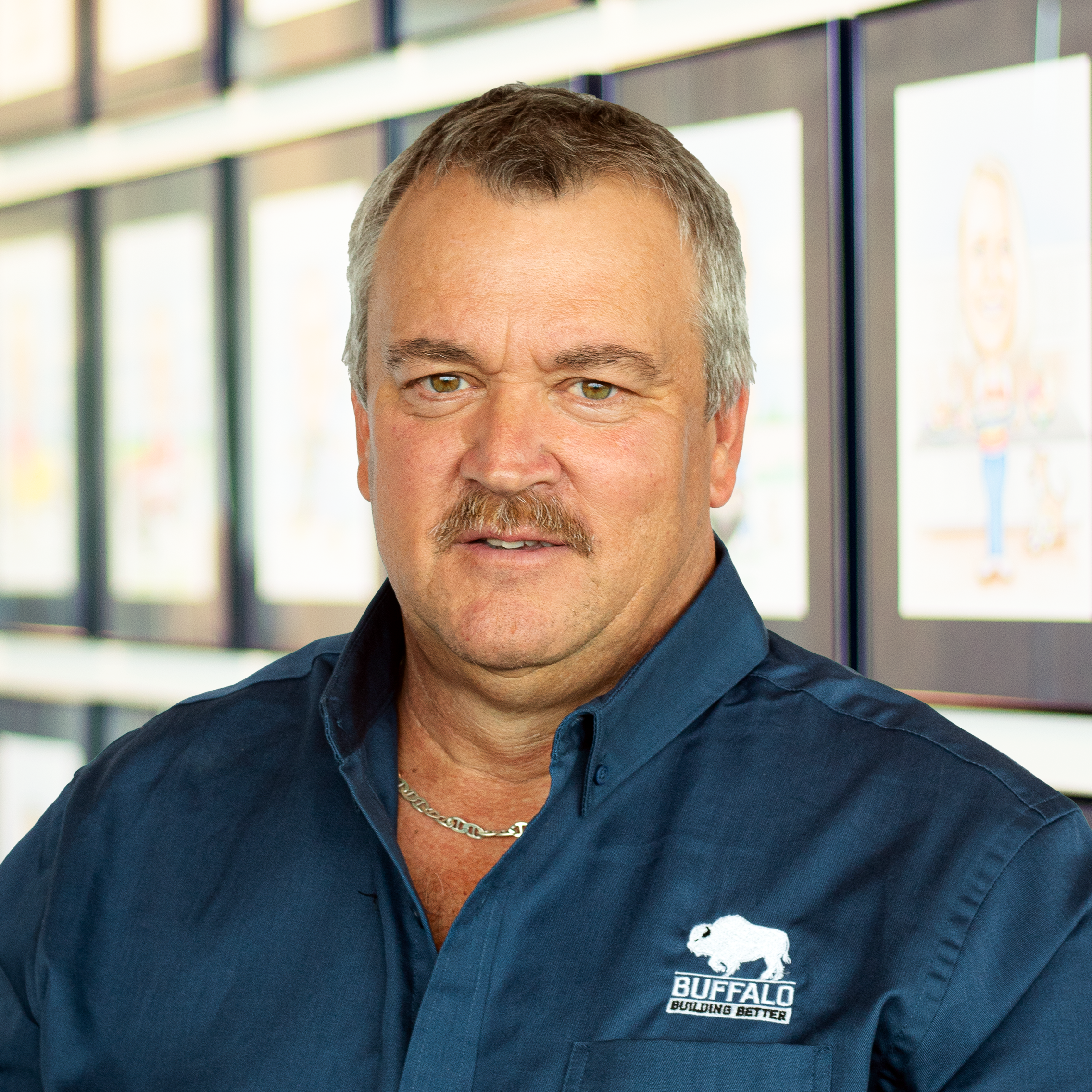 Steve Fordenbacher wearing a buffalo branded button down shirt, while standing in front of a wall displaying framed caricatures.