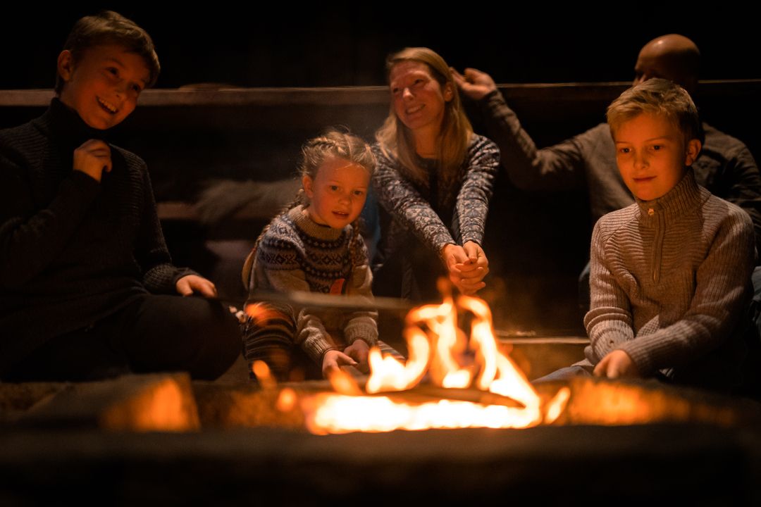 A family is sitting around a campfire pan. They are smiling and wearing wool jumpers