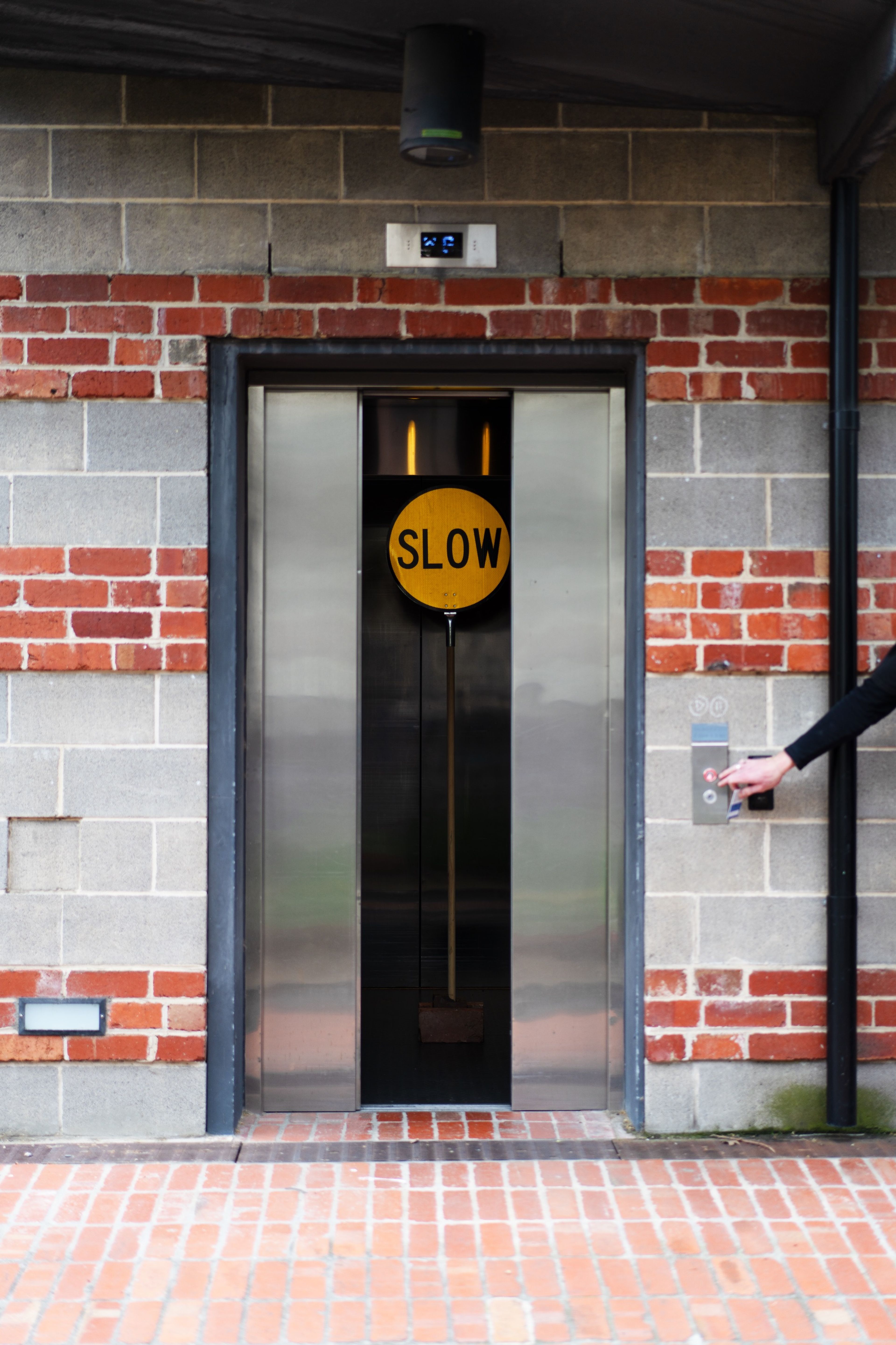 A yellow "SLOW" sign is set in the open doors of an elevator against a modern industrial background. A hand reaches for the elevator button, adding an interactive element to the installation.