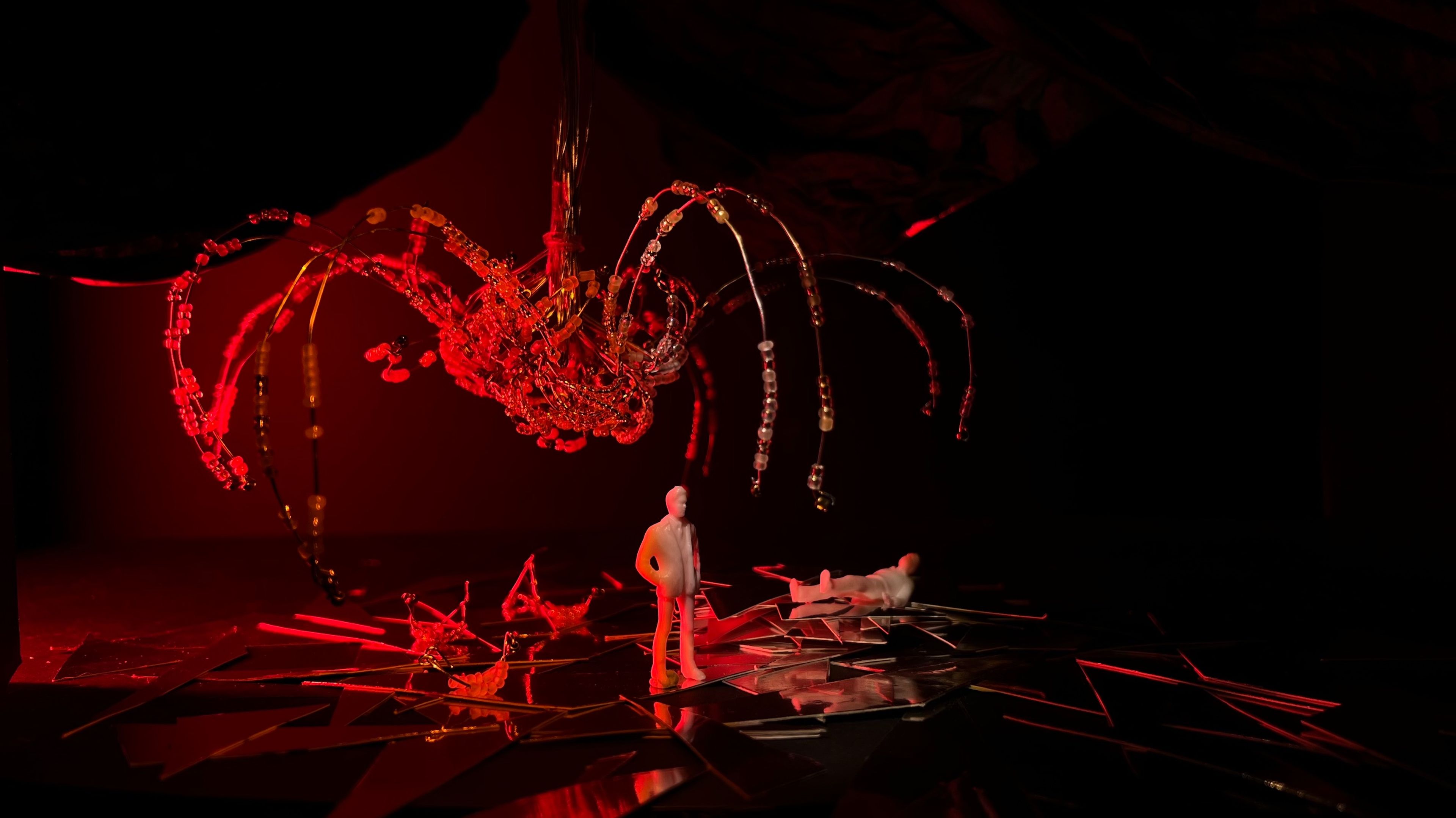 Miniature set design illuminated in red, showing a white figure on a fractured reflective surface beneath an arching beaded chandelier structure