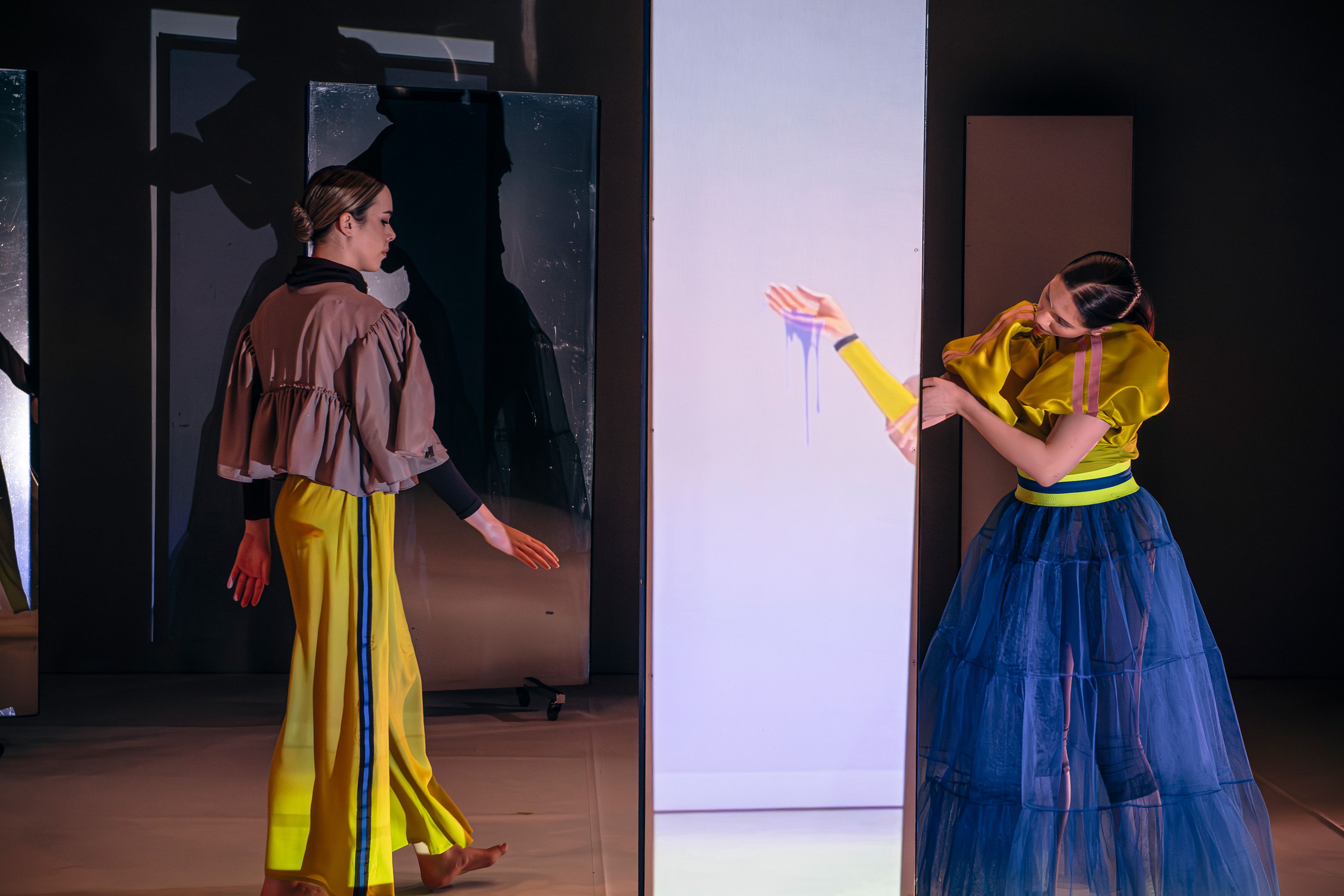 Dance production photograph of two dancers facing each other, framed by mirrored panels, with one dancer extending her arm while gazing toward her reflection.