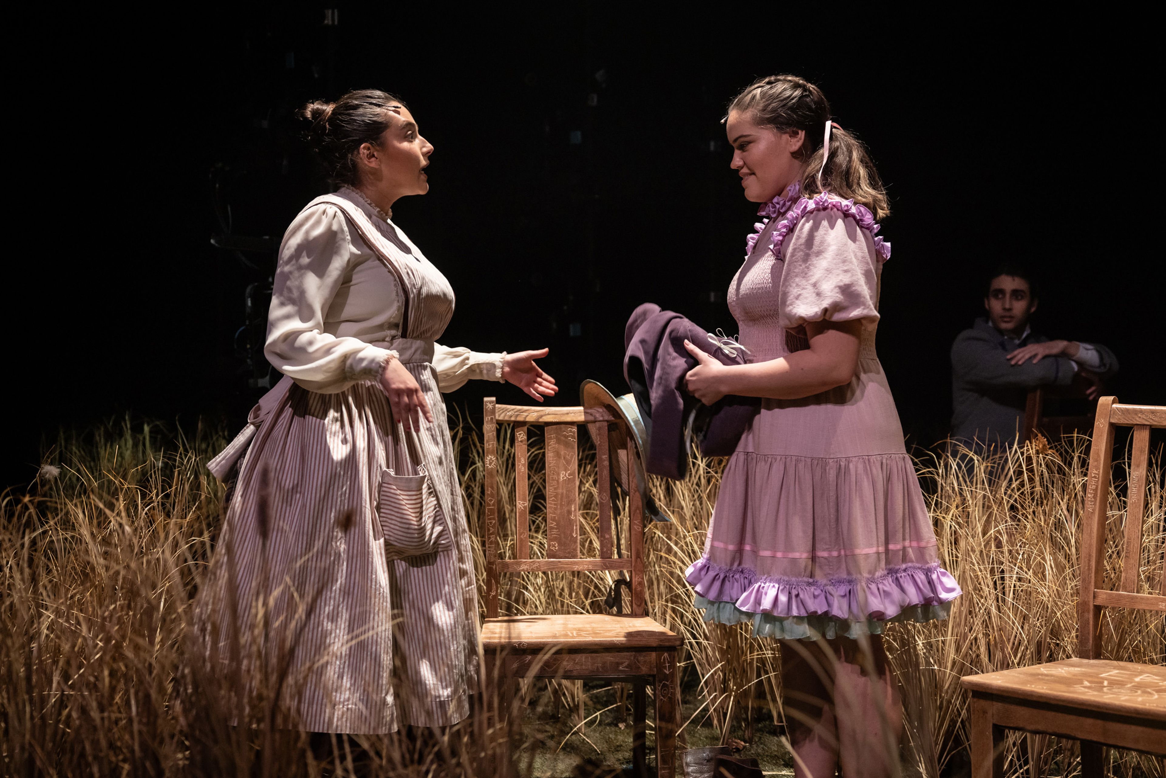 Theatre production photograph showing two performers in Victorian-era dresses having an intense conversation amongst wooden chairs and wheat grass, with a figure partially visible in the background.