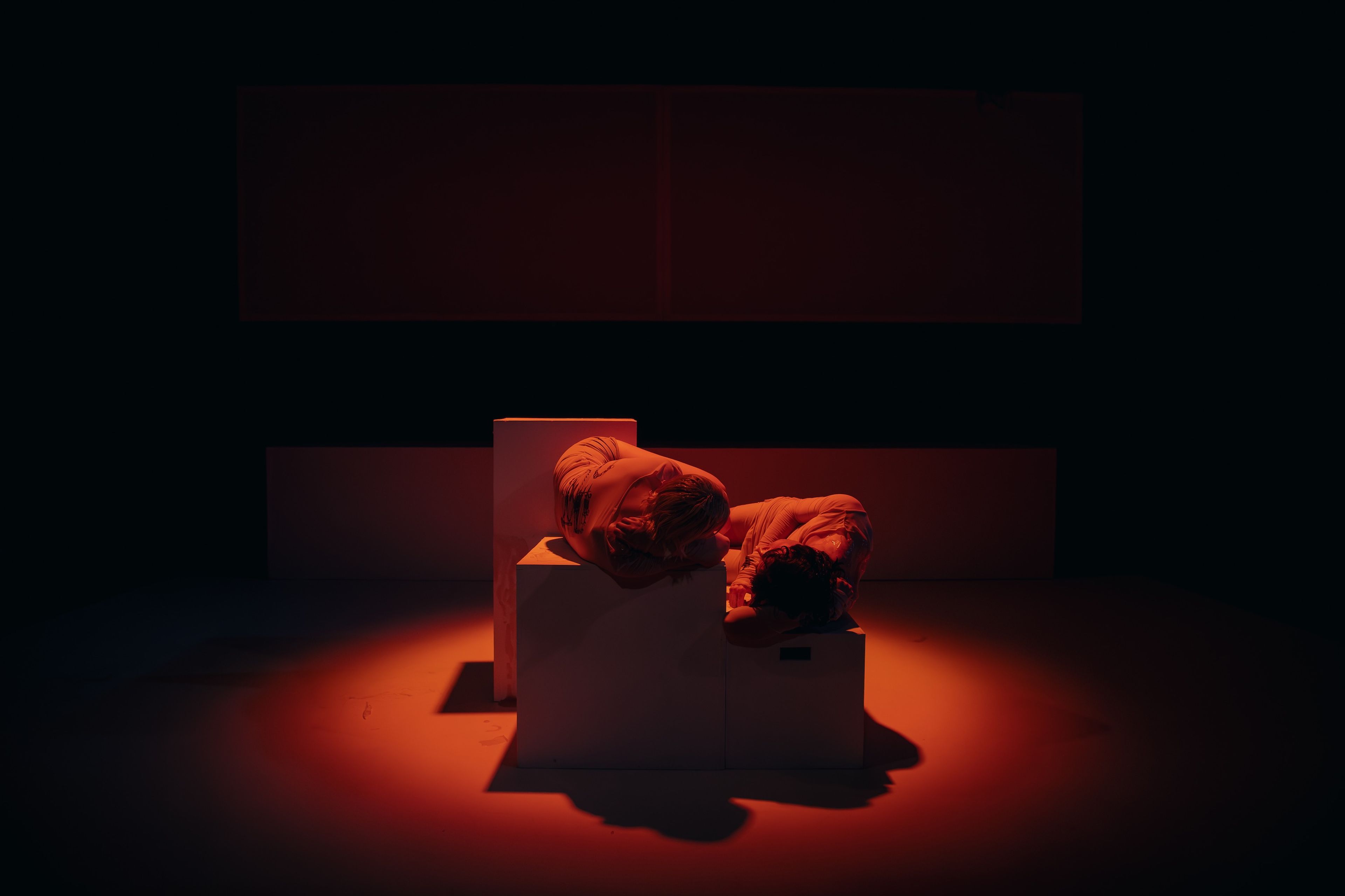 Performance photograph of two figures resting their heads on white plinths, dramatically lit in deep red lighting against a black background