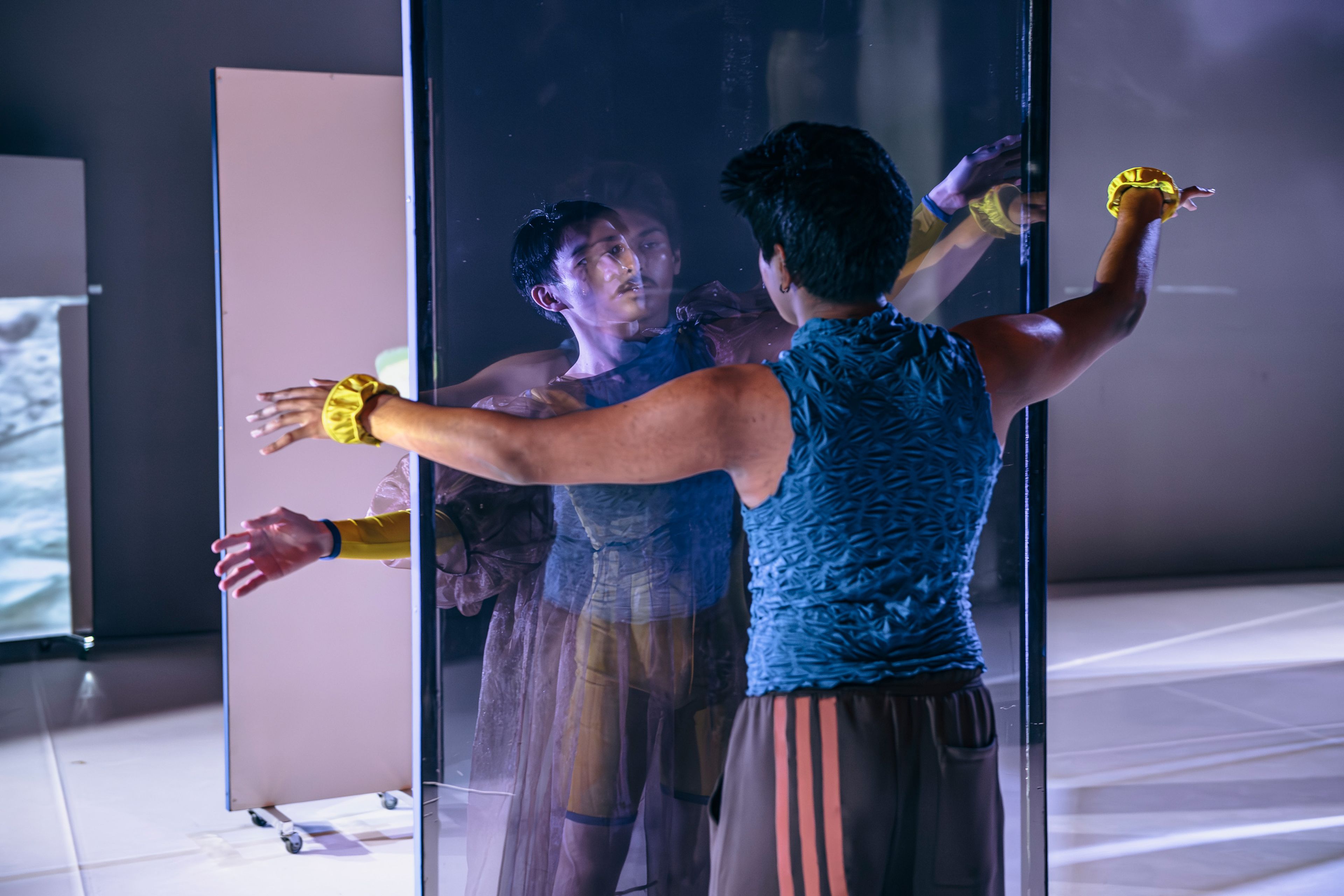 Dance production photograph of a dancer pressing against a mirrored panel, with reflections and shadows creating a sense of duality.