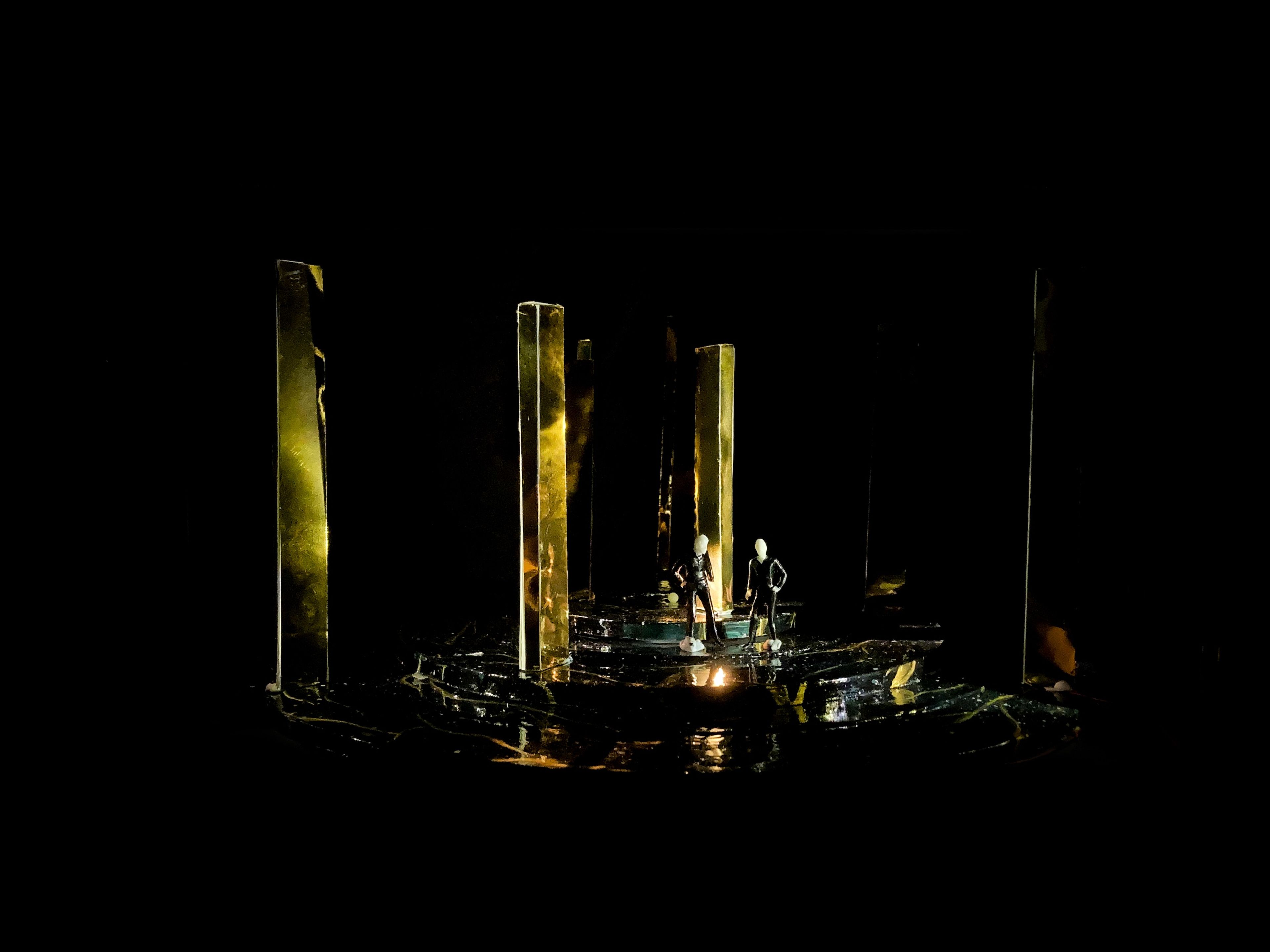 Stage model for Act 2, Scene 4, with two small figures surrounded by tall golden panels in a dimly lit space.