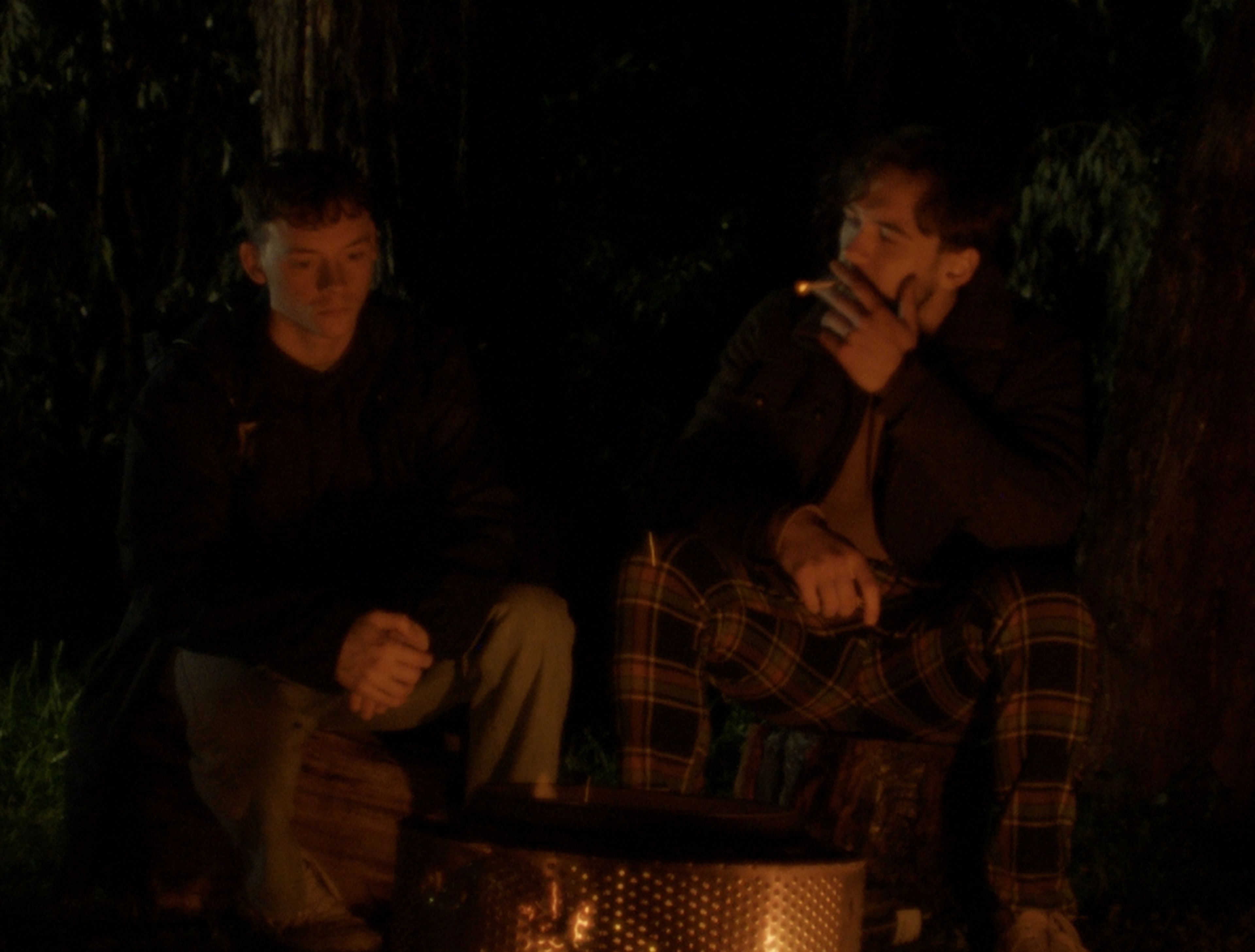 Film still capturing two young men sitting outdoors at night, one in plaid pyjamas, lit dramatically by the warm glow of a fire pit between them.