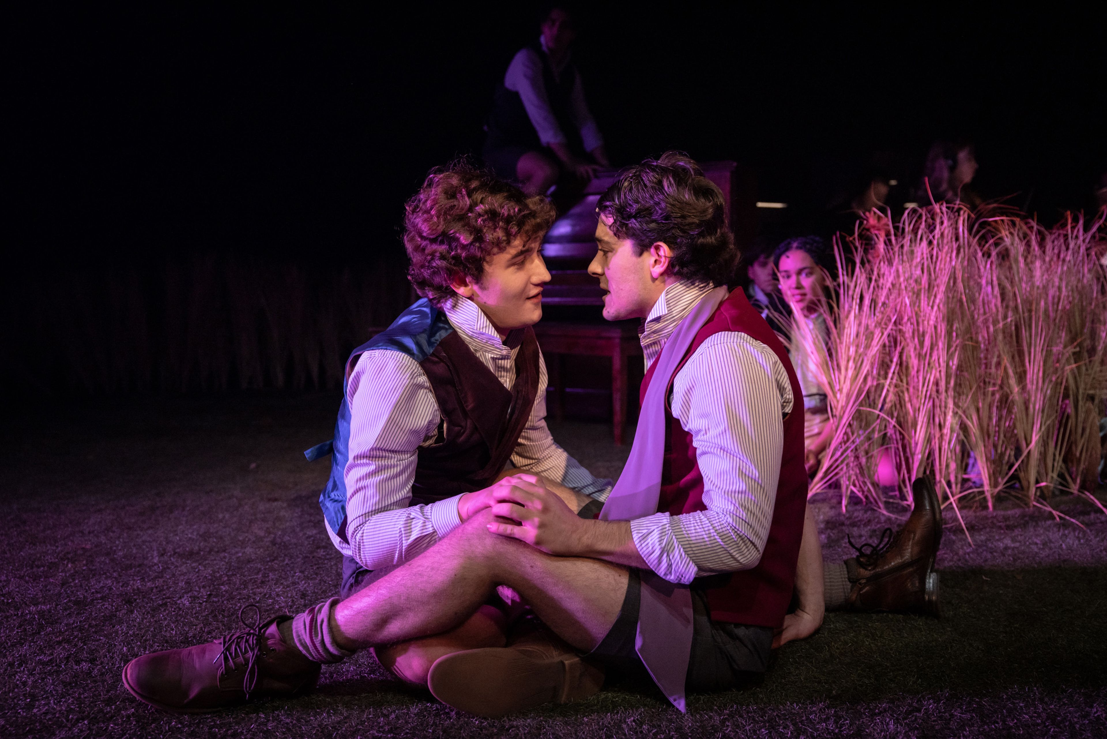 Theatre production photograph capturing an intimate moment between two male performers in period dress with striped shirts and waistcoats, seated amongst wheat grass with purple theatrical lighting.