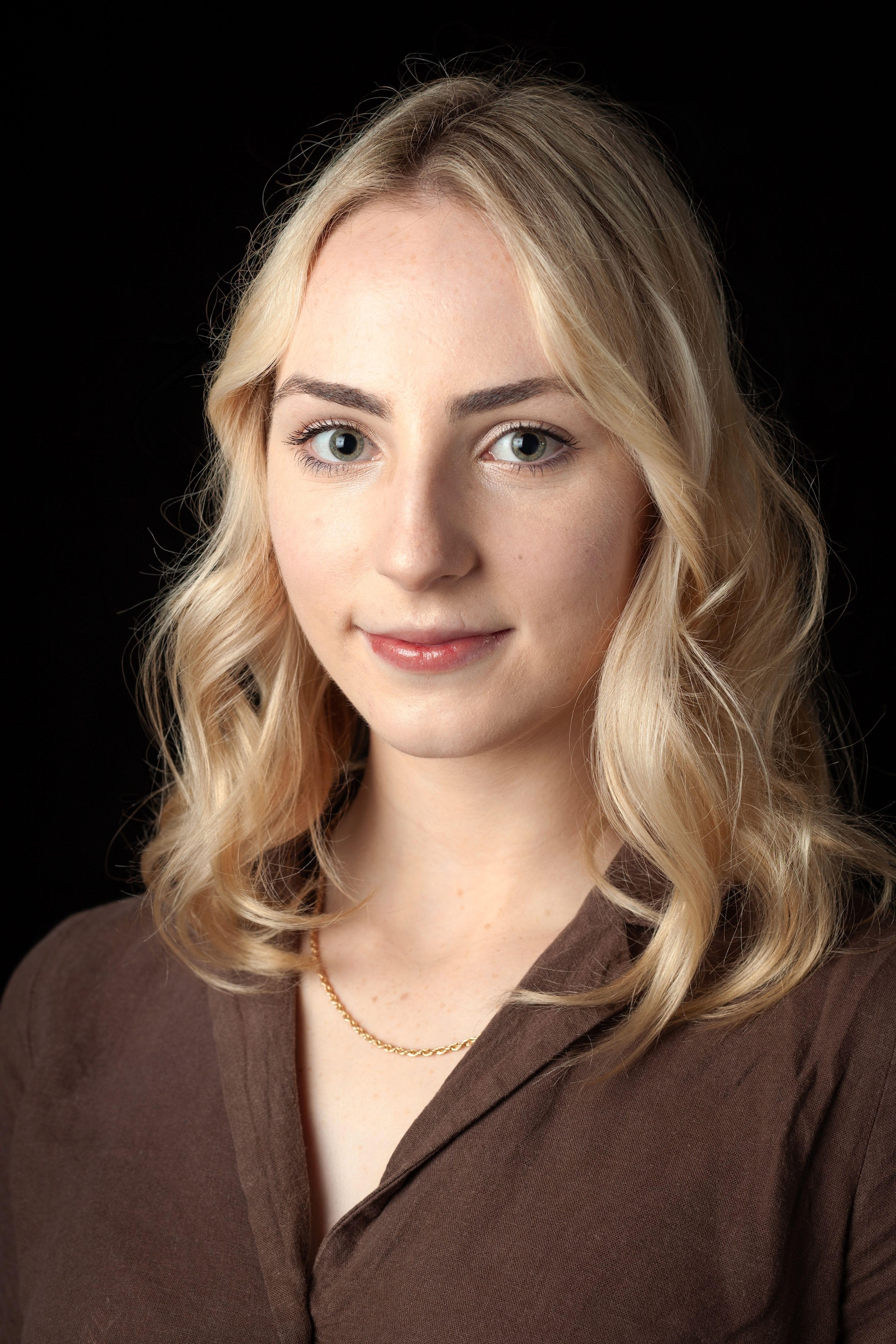 Headshot of a young woman with wavy blonde hair, wearing a brown shirt and a gold necklace, against a black background.