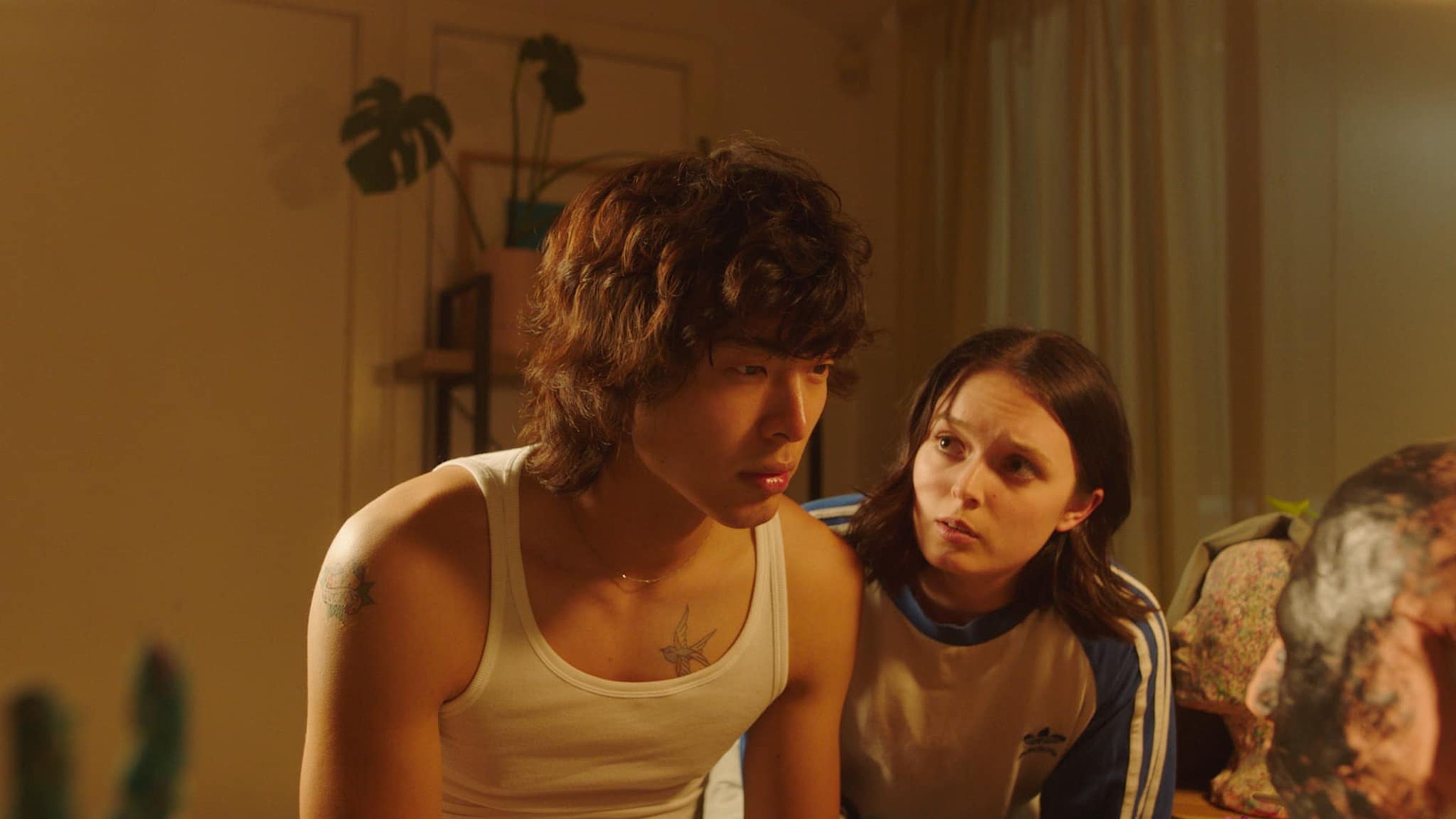 Film still of a young man in a white tank top sitting closely with a woman who looks at him with concern in a softly lit room.