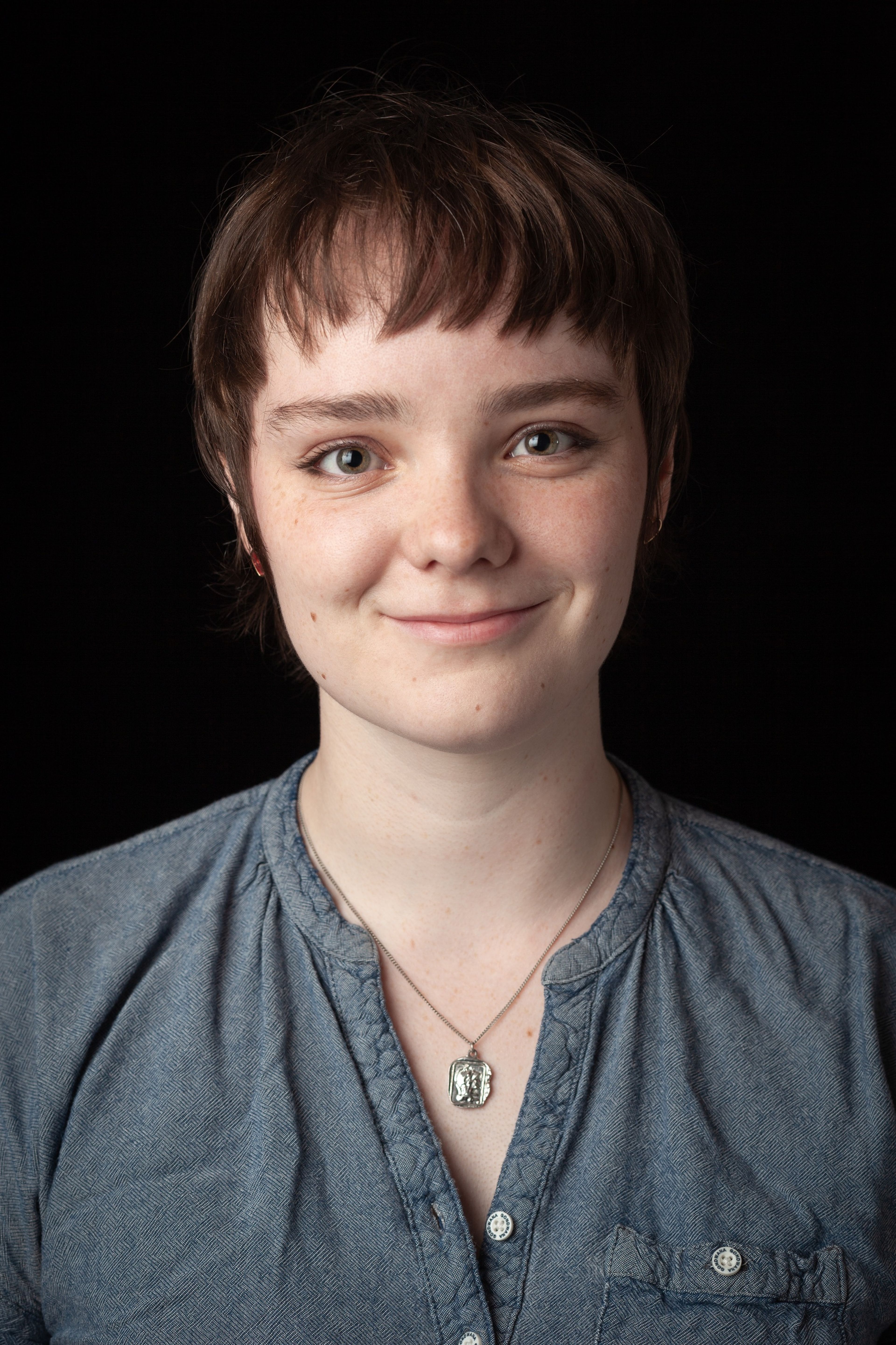 Headshot of Viv, who has short dark hair, and is wearing a blue button-up shirt and a pendant necklace, against a black background.