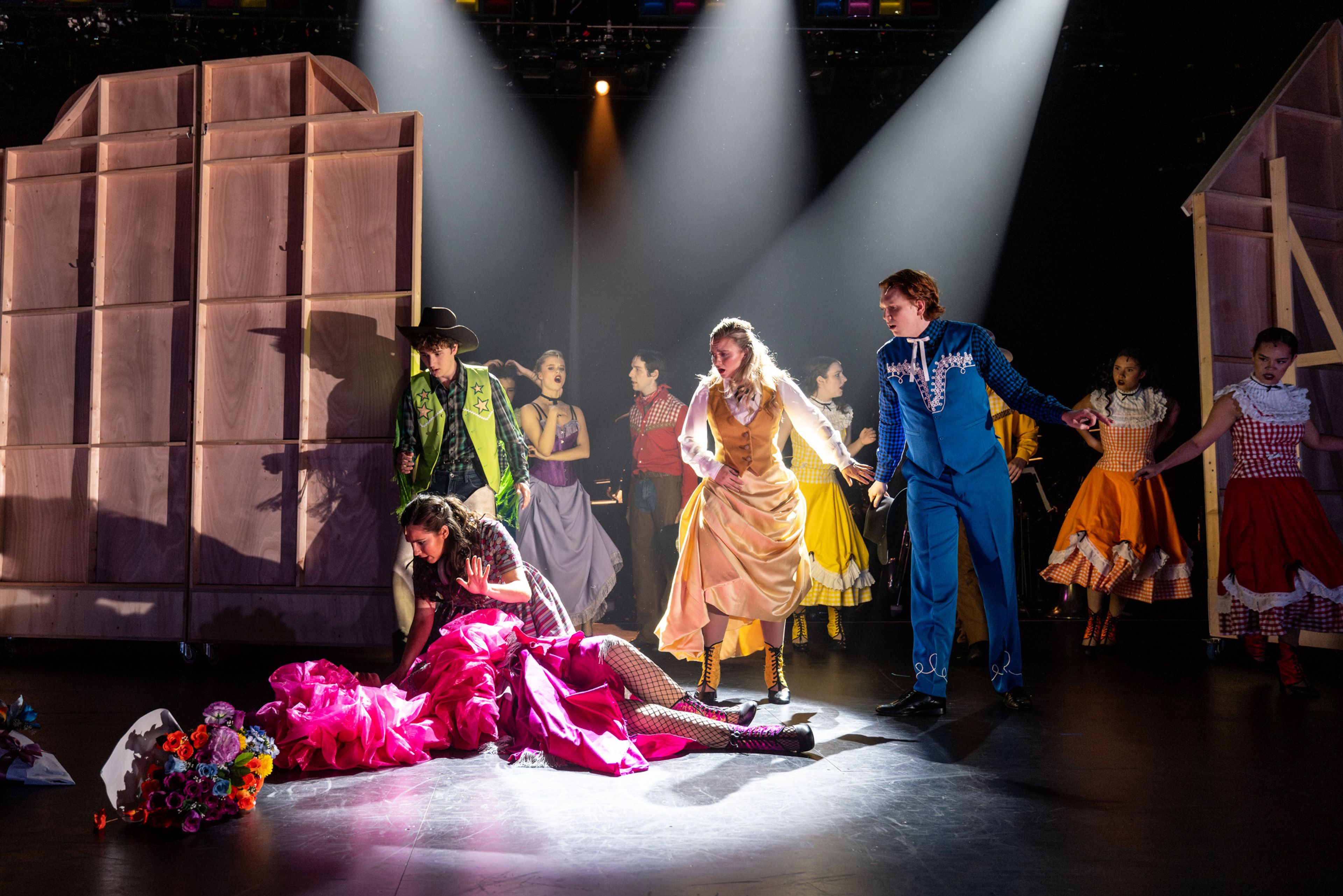 Theatre production photograph of a dramatic scene with a performer in a bright pink costume on the floor, surrounded by other cast members in Western attire under focused spotlights.