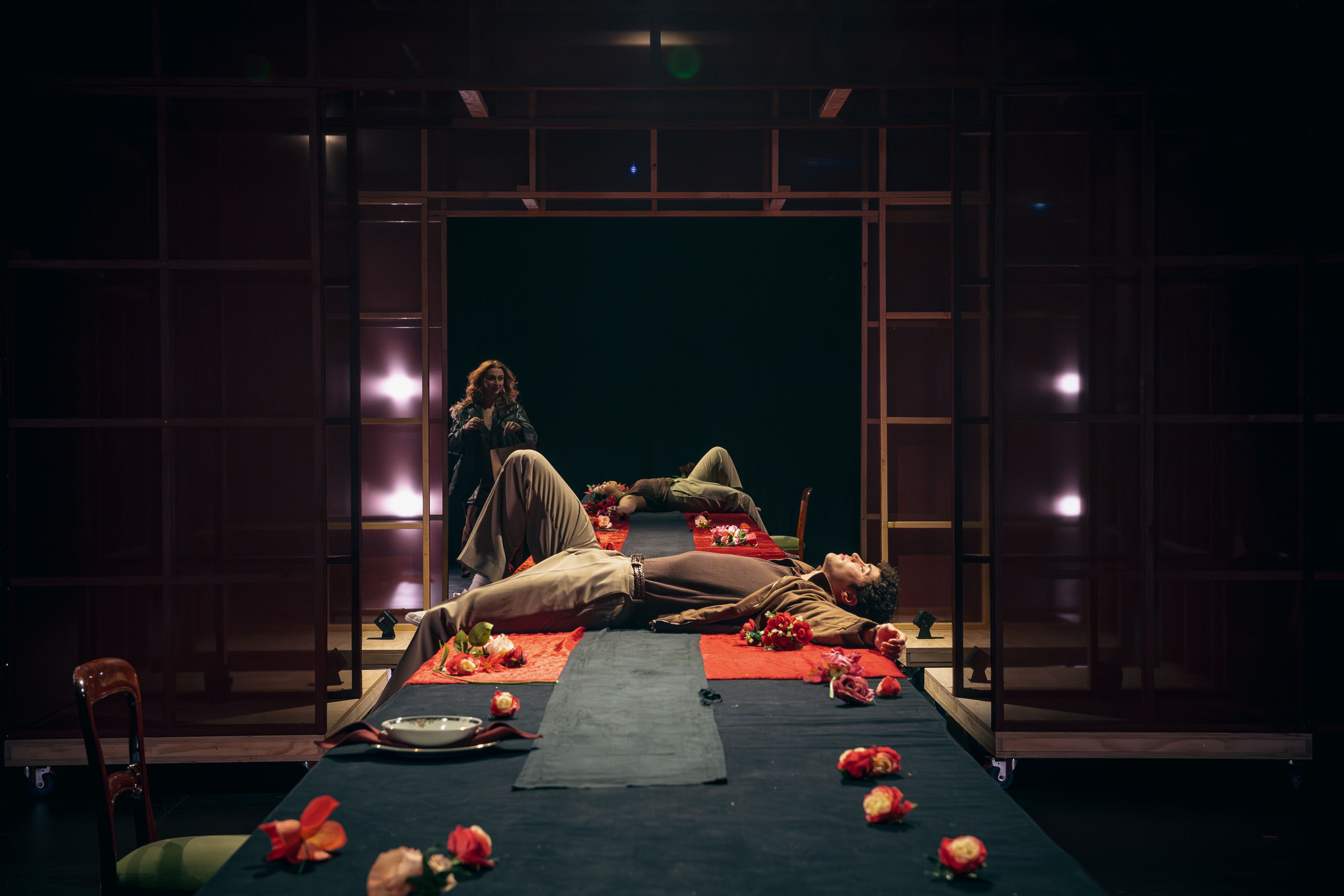 Theatre production photograph showing actors lying on a long table decorated with red flowers, while another actor stands in the background, framed by a wooden structure.