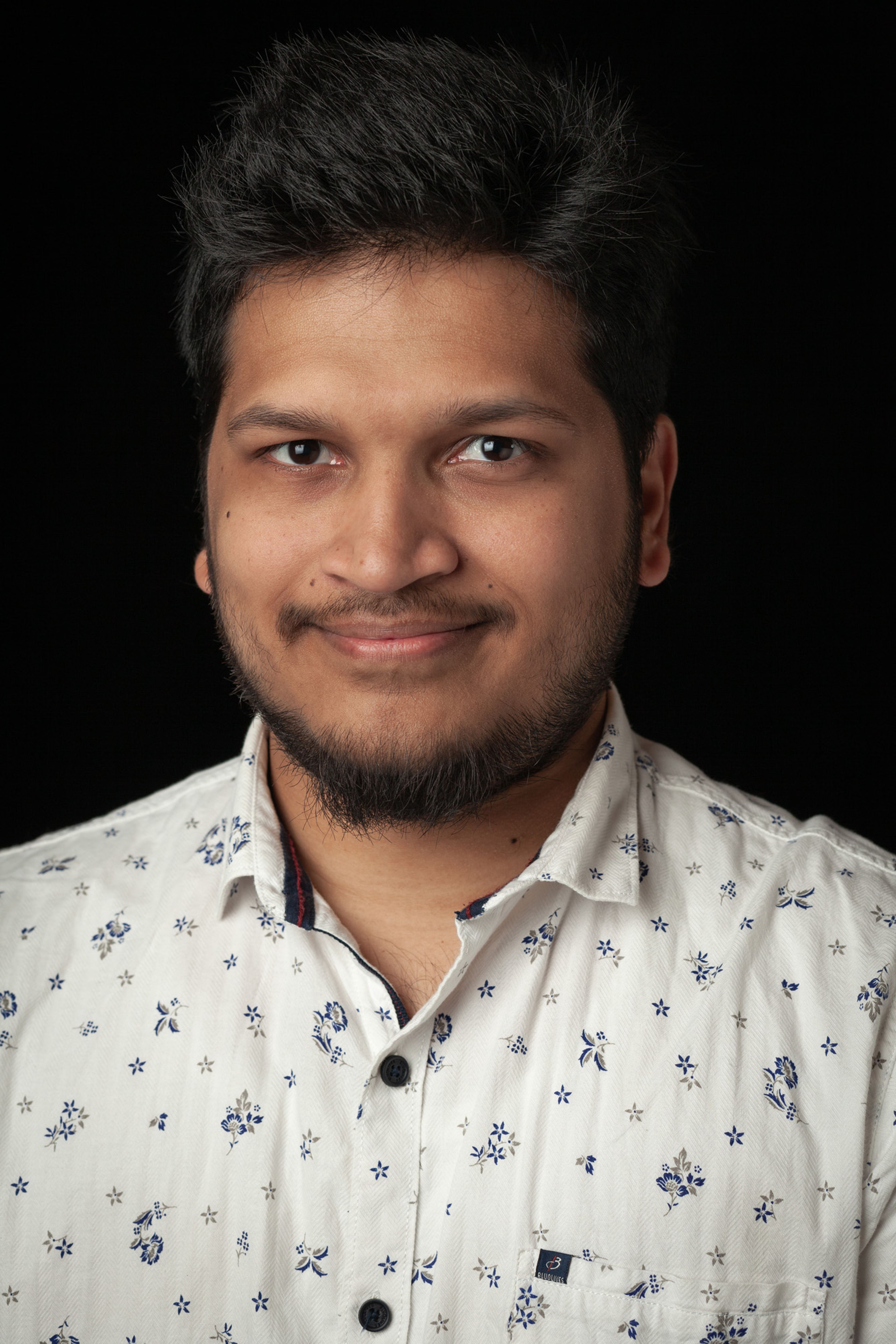 Headshot of a man with short, dark hair and a trimmed beard, wearing a white, patterned button-up shirt with small blue floral designs, set against a black background.