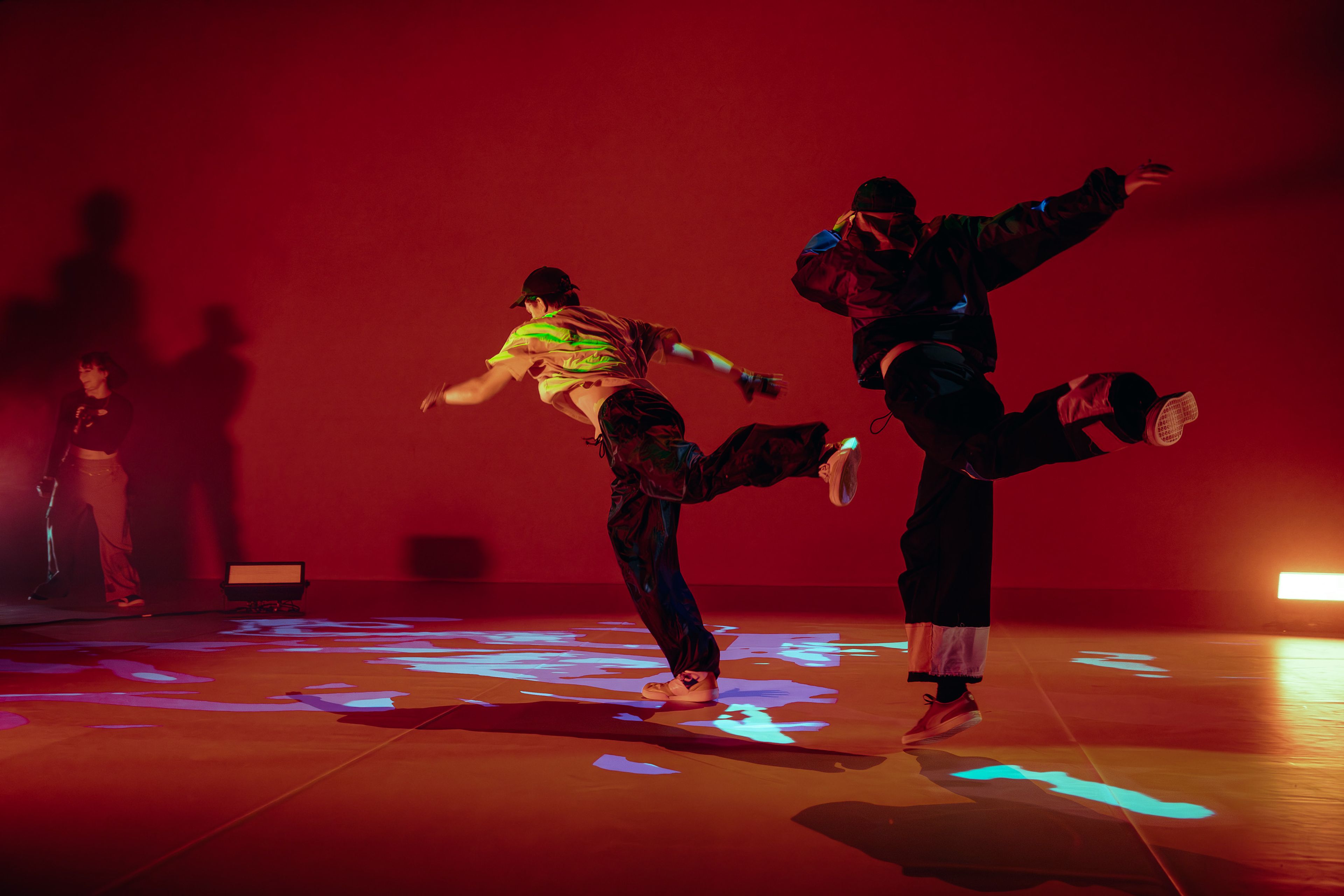 Dance production photograph of two dancers leaping in unison, casting shadows on a vivid red stage floor, with colourful lighting effects projected beneath them.