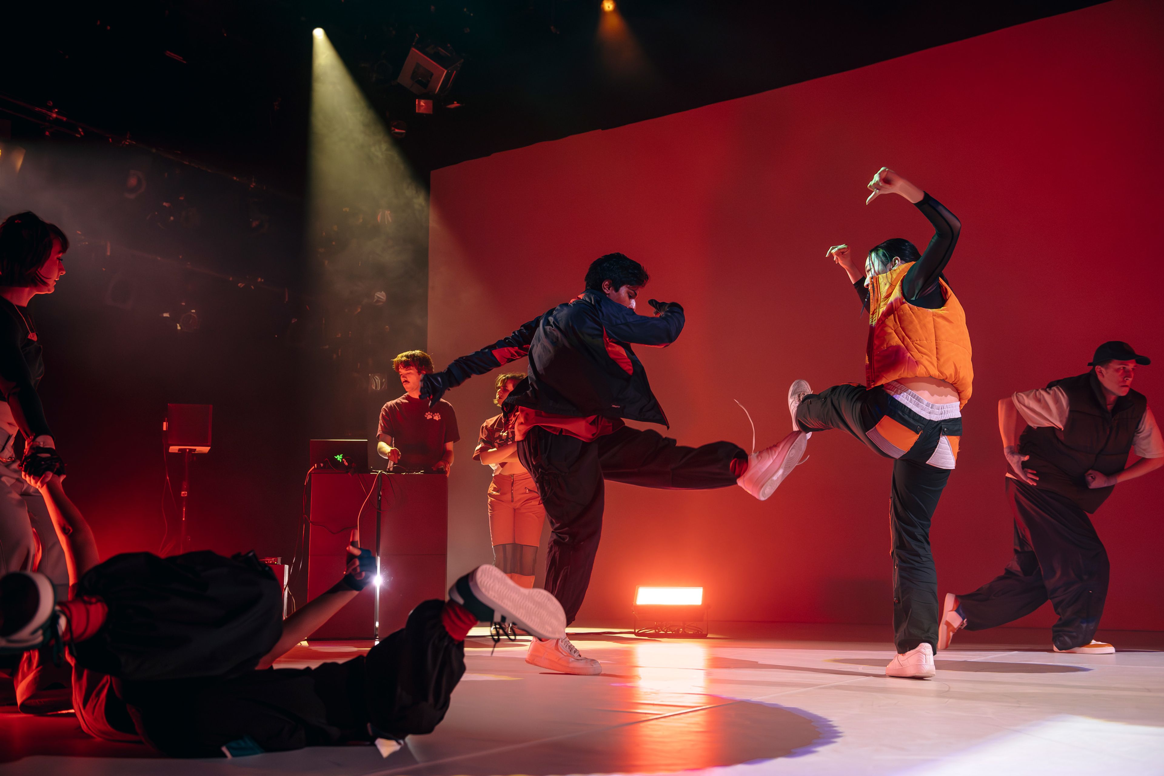 Dance production photograph of dancers in motion, one performing a high kick, against a vibrant red backdrop with dynamic stage lighting.