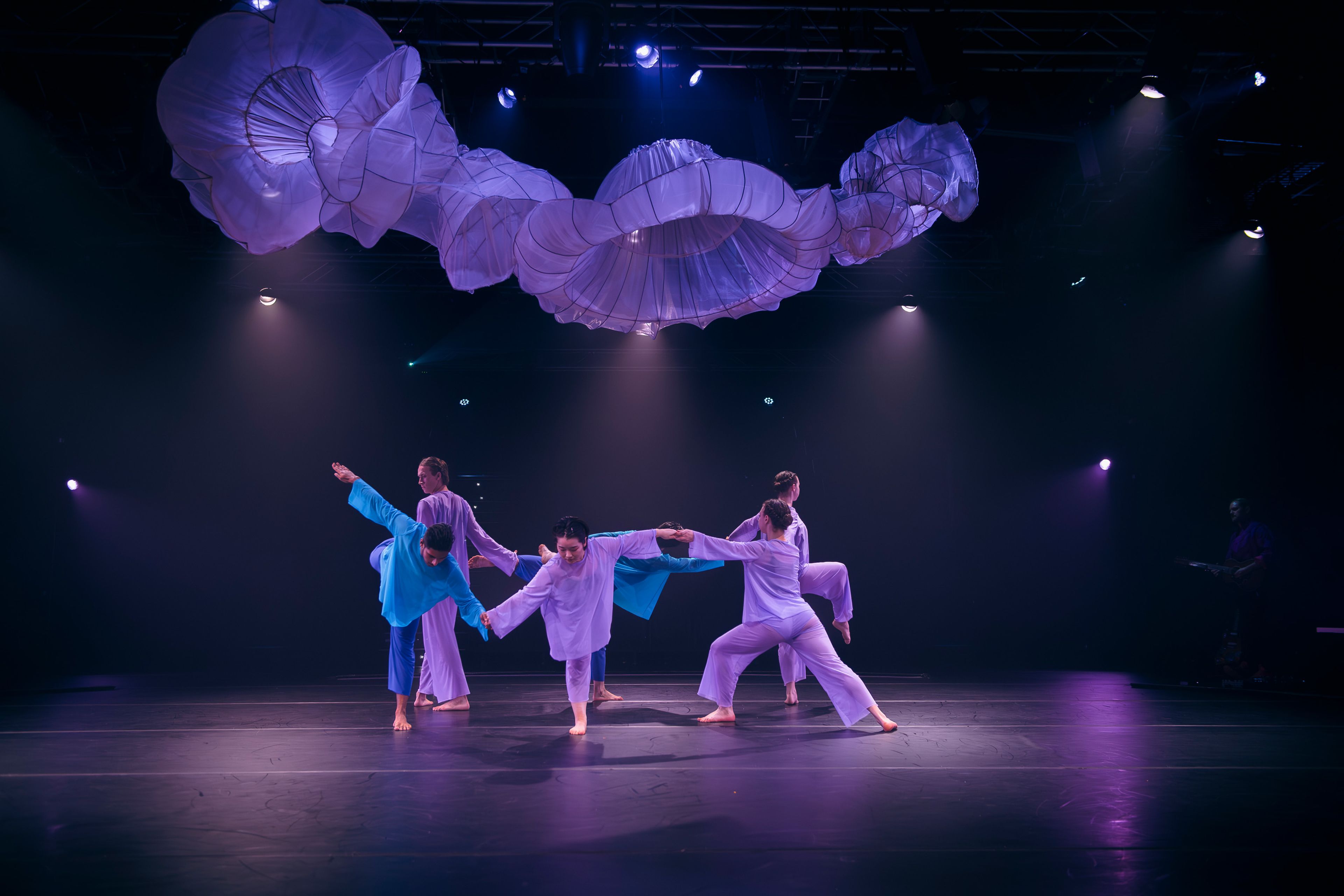 Theatre production photograph showing a group of dancers in pastel-coloured costumes performing a balanced pose, with sculptural white fabric forms suspended above and purple lighting creating an ethereal atmosphere.