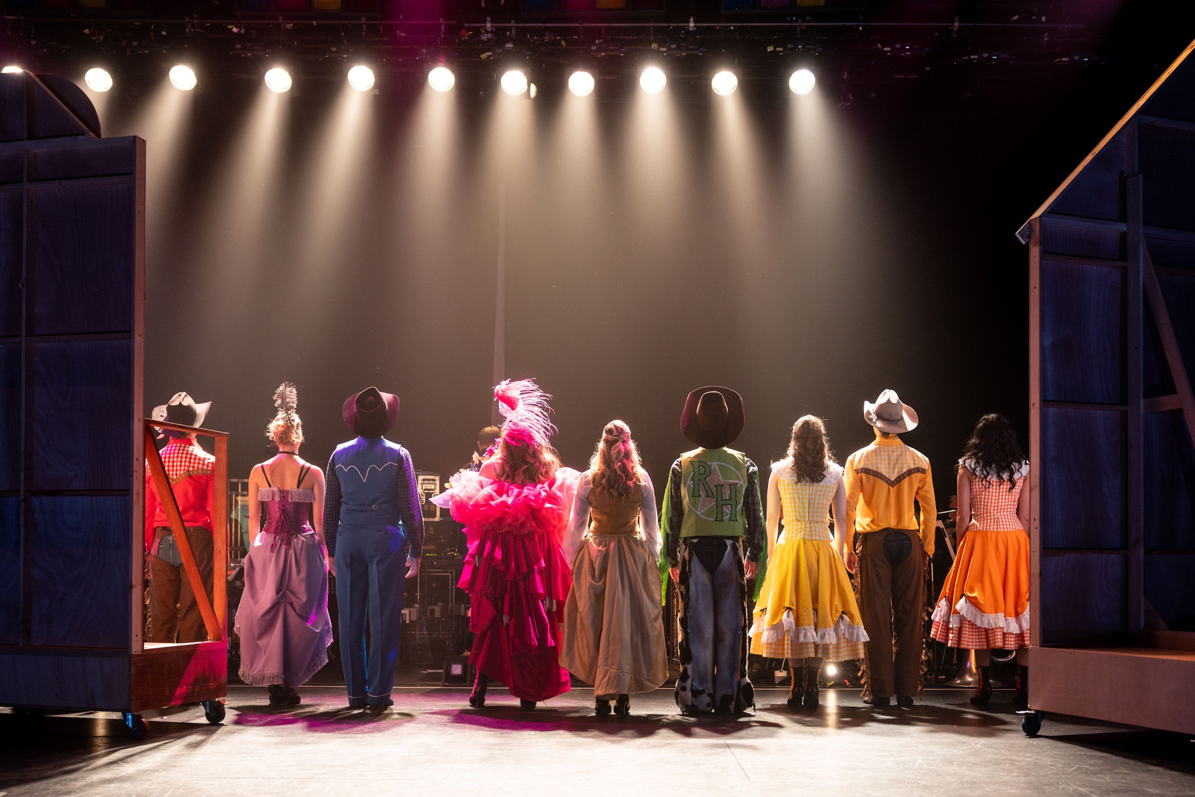 Theatre production photograph showing performers dressed in colourful western-themed costumes standing in a line facing away from the camera, with bright overhead lights illuminating the stage.