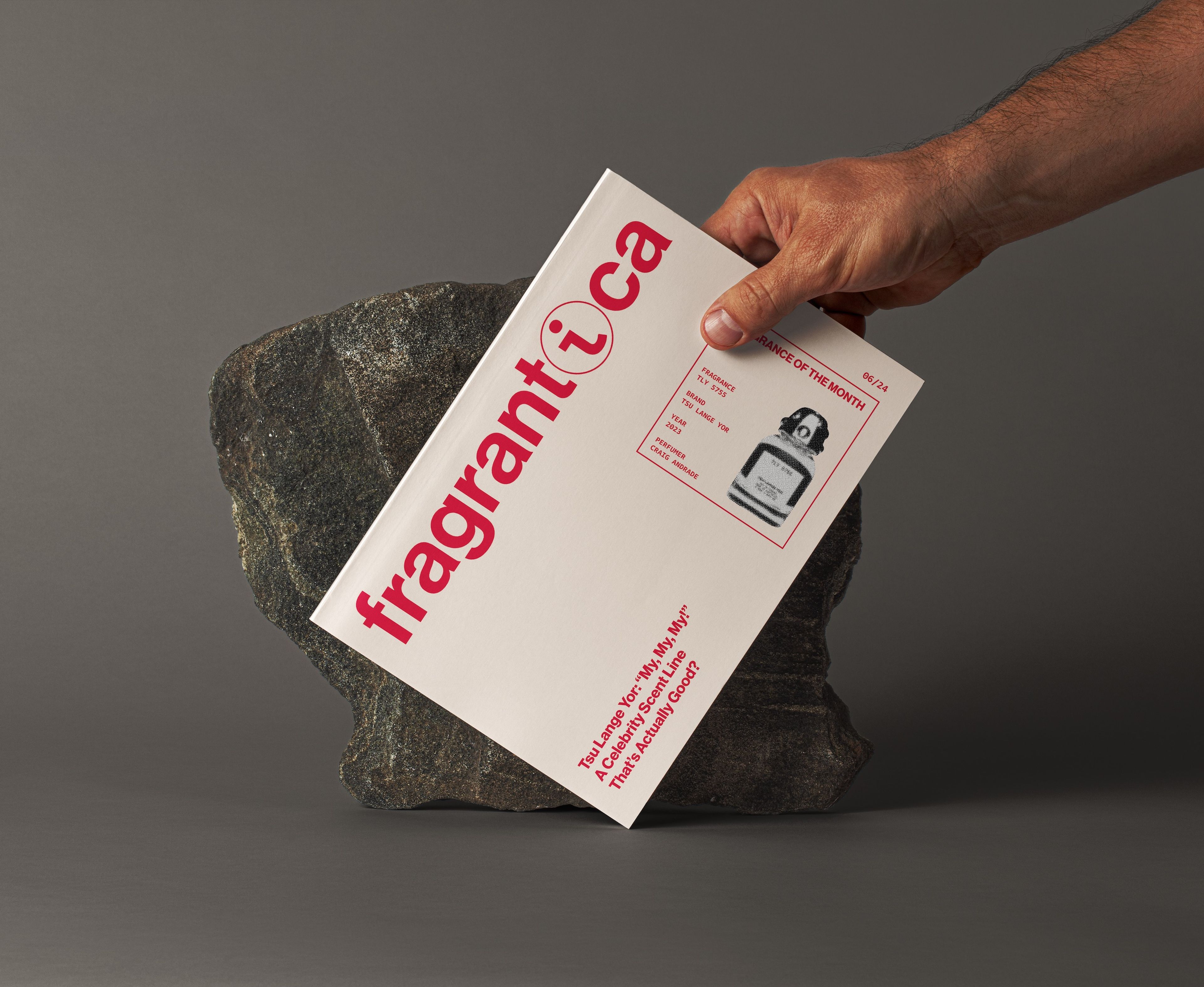 A hand holds a Fragrantica magazine against a textured stone, with the cover displaying the red Fragrantica logo and a feature on the "Fragrance of the Month.