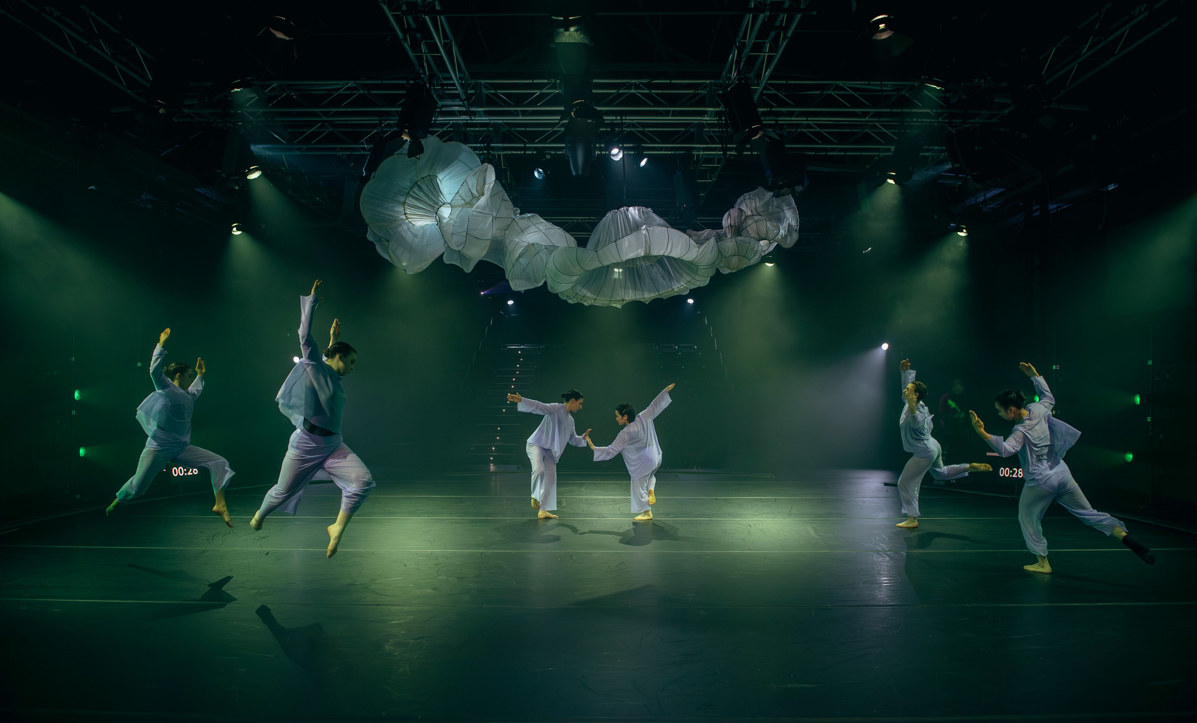 Theatre production photograph showing dancers in white costumes performing synchronised movements across the stage, with sculptural white fabric forms suspended above and green-tinted lighting creating atmosphere.