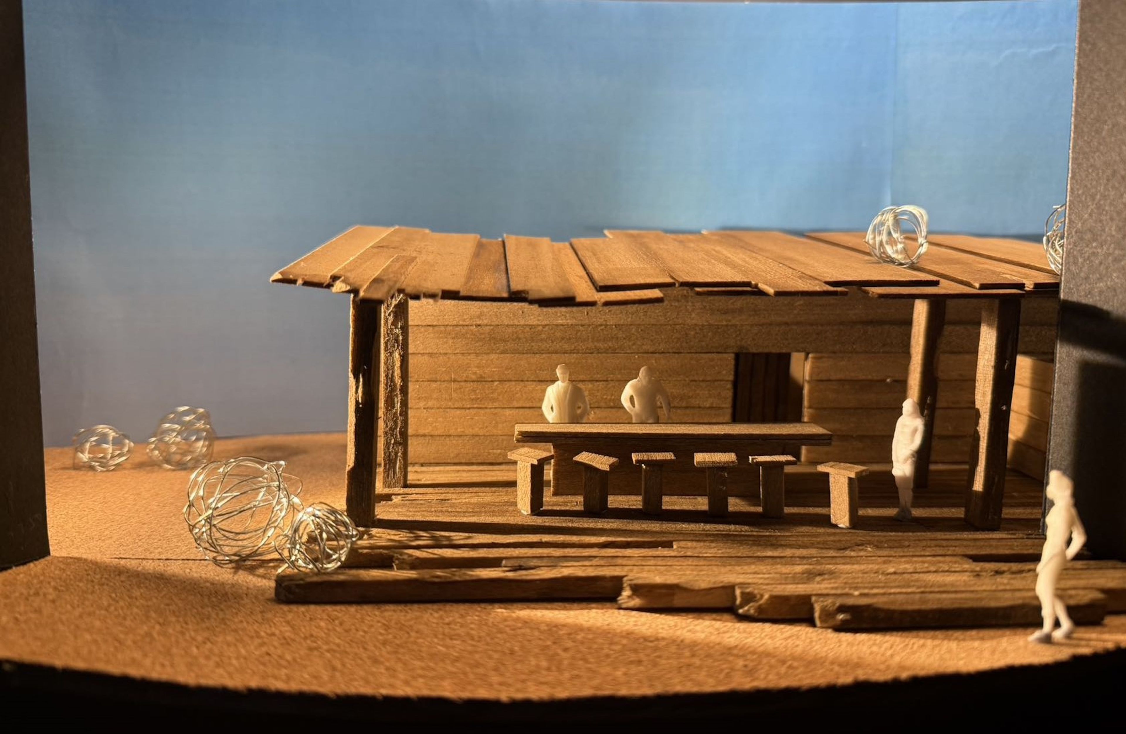 Scale model of a saloon set design during daylight, with two figures at a table inside and wire tumbleweeds scattered outside.