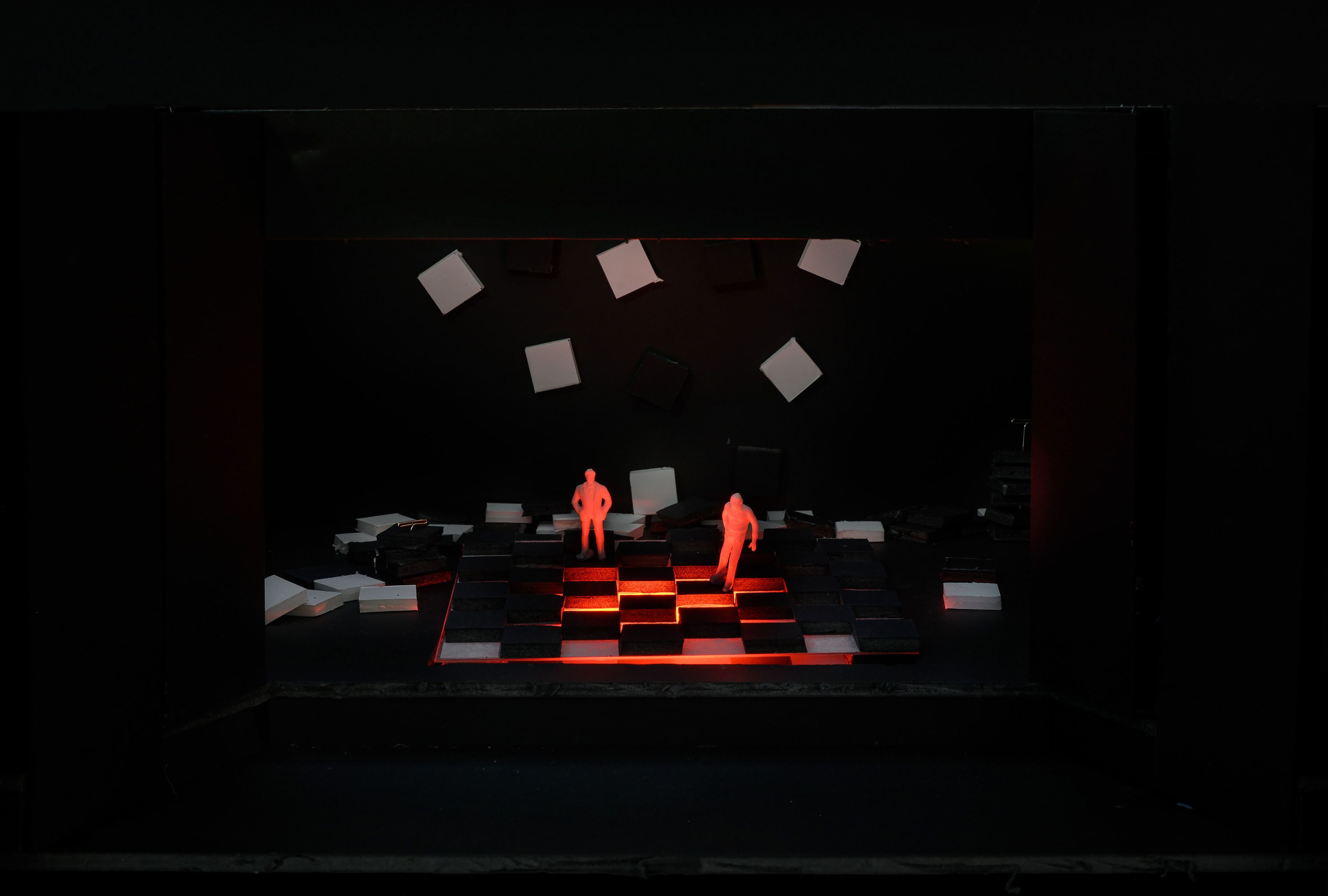 A theatrical stage model with a checkerboard floor illuminated by red light, creating a dramatic and eerie ambiance. Two small human-like figures stand on the stage, surrounded by scattered black and white squares suspended and scattered around the space