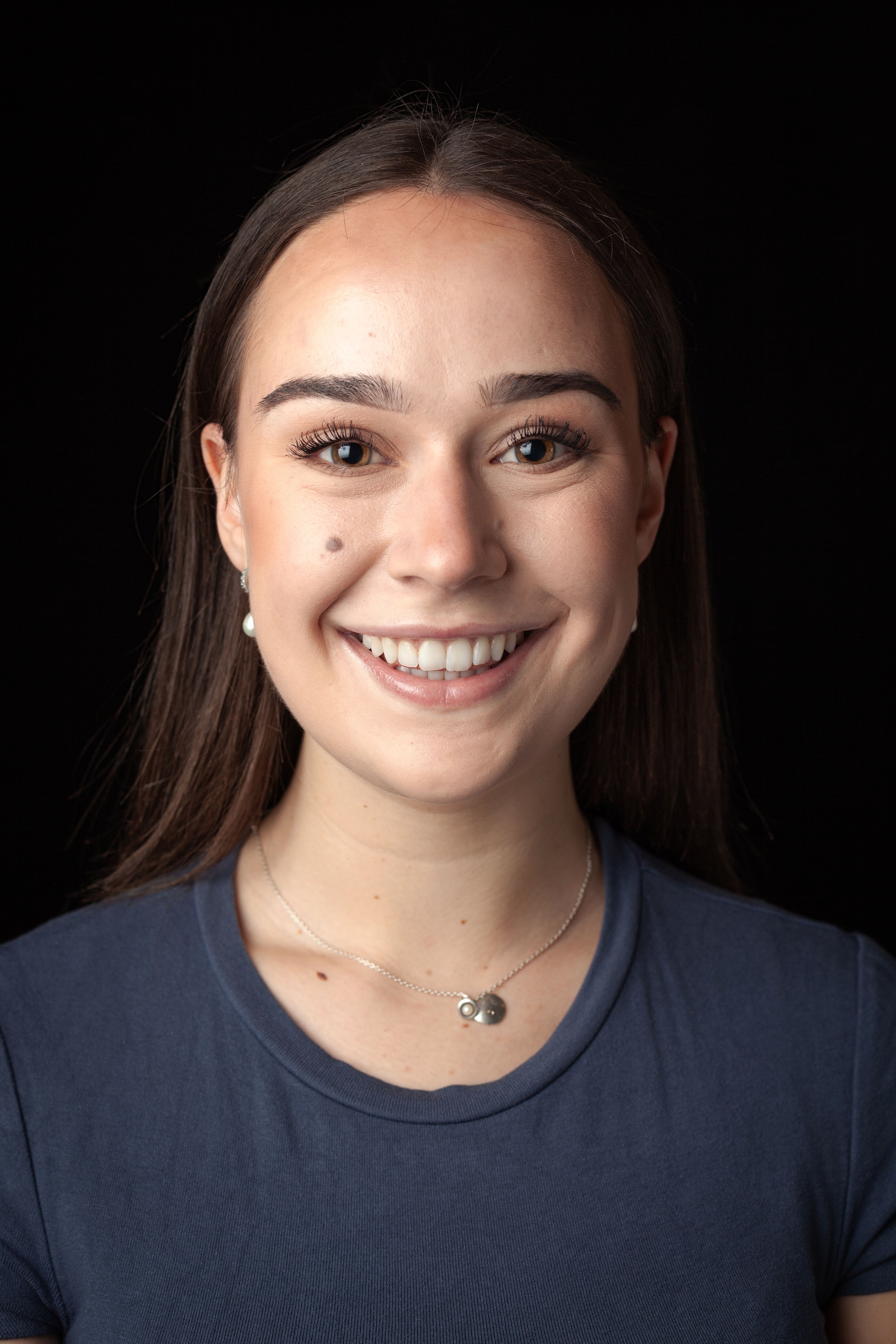 Headshot of a smiling young woman with straight brown hair, wearing a blue top and a delicate necklace with a silver pendant, against a black background.