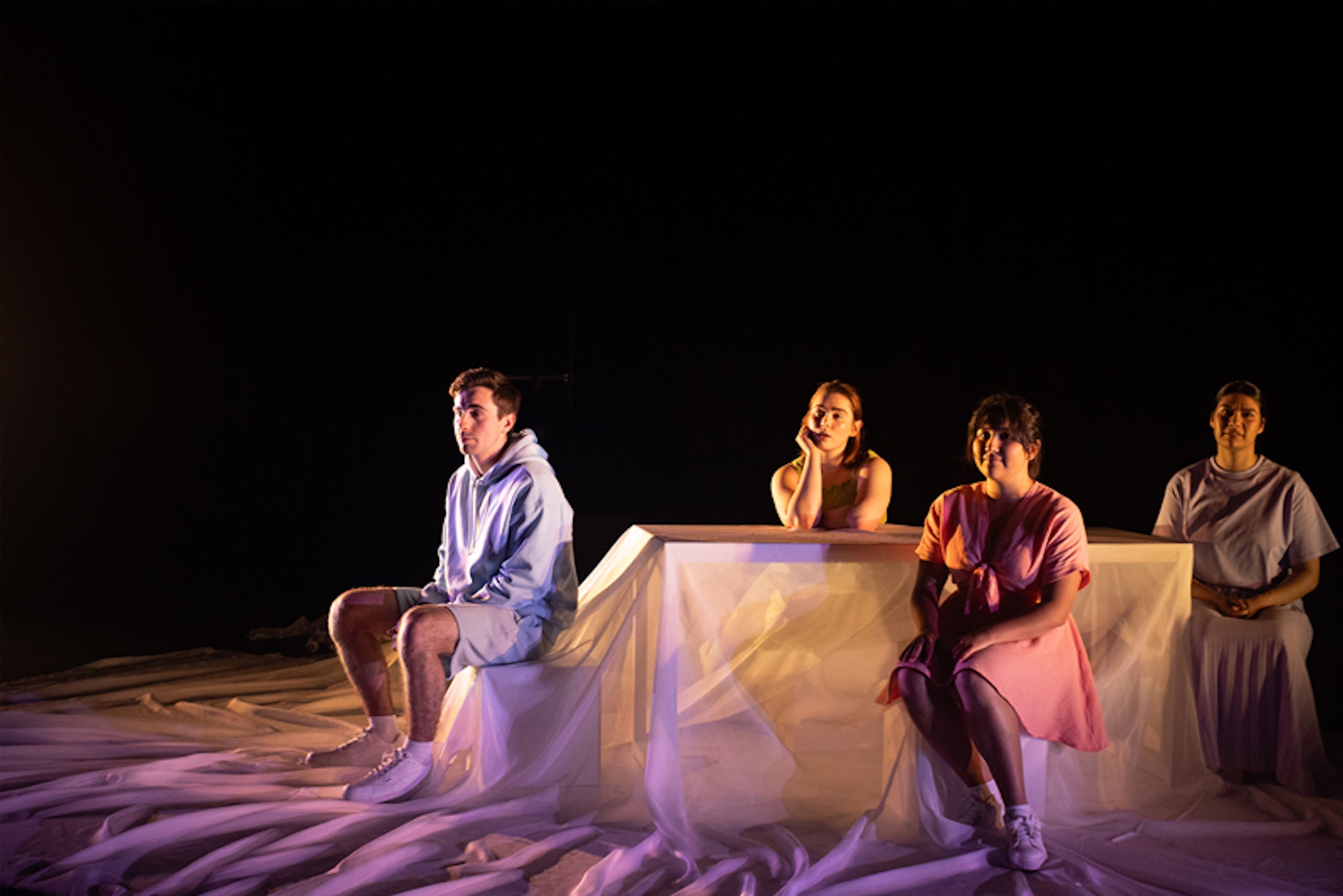 Production photograph of four performers on a minimalist white set, lit with purple lighting, wearing casual contemporary clothing while seated at different heights