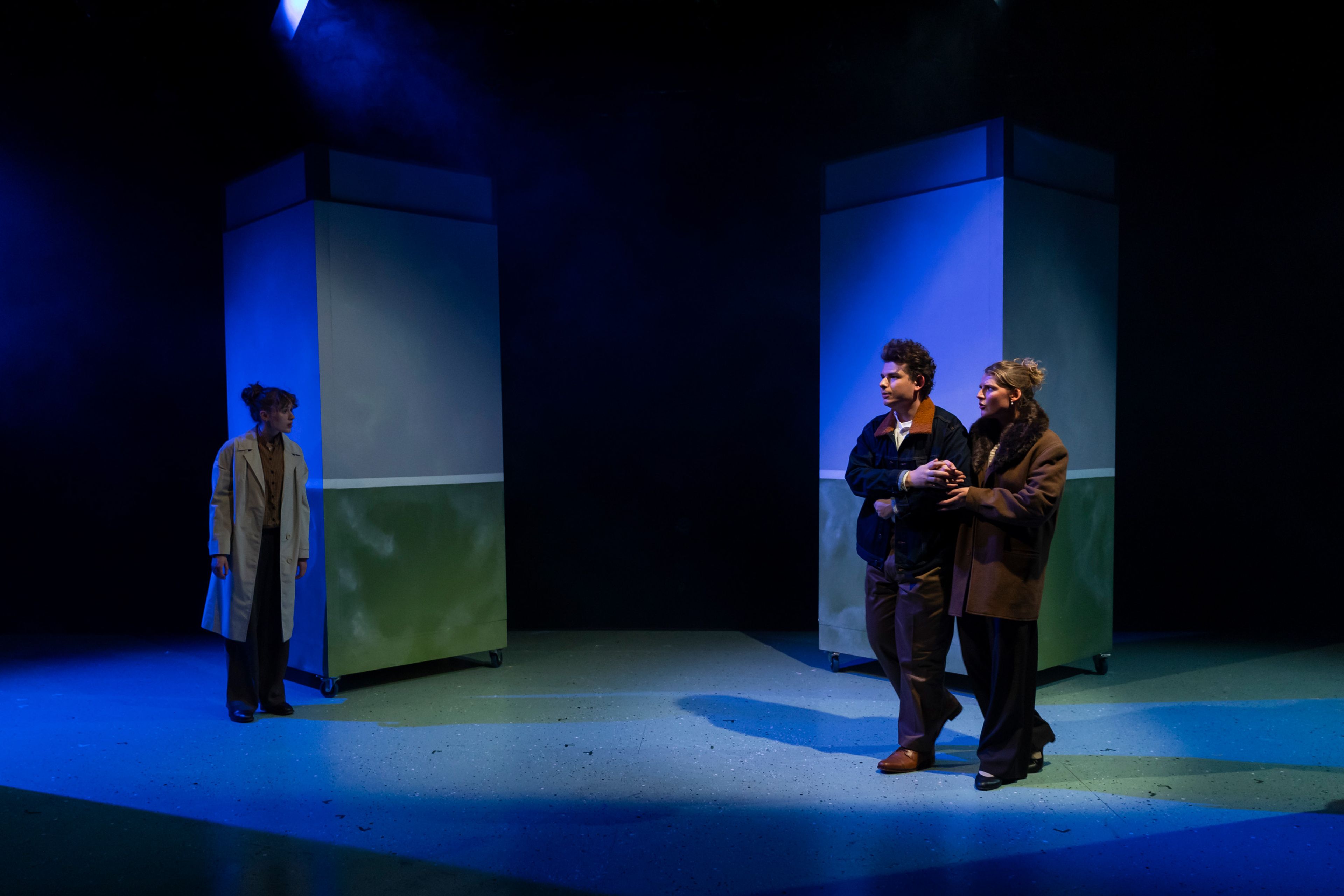 Theatre production still featuring three performers in winter coats arranged around minimalist set pieces with blue atmospheric lighting