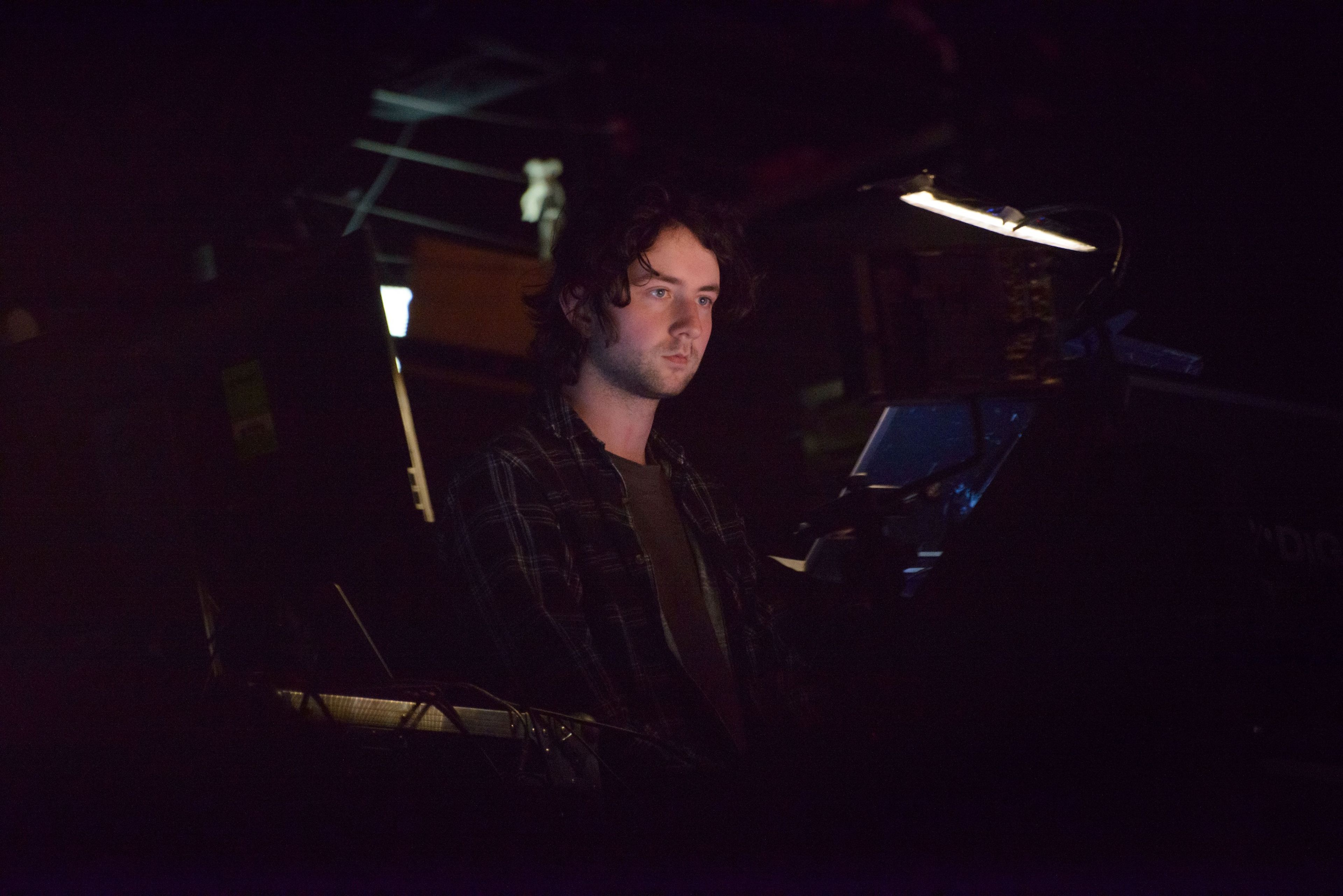 A person with curly hair sits in a dimly lit control booth, focused on a screen in front of them, with various equipment surrounding them.