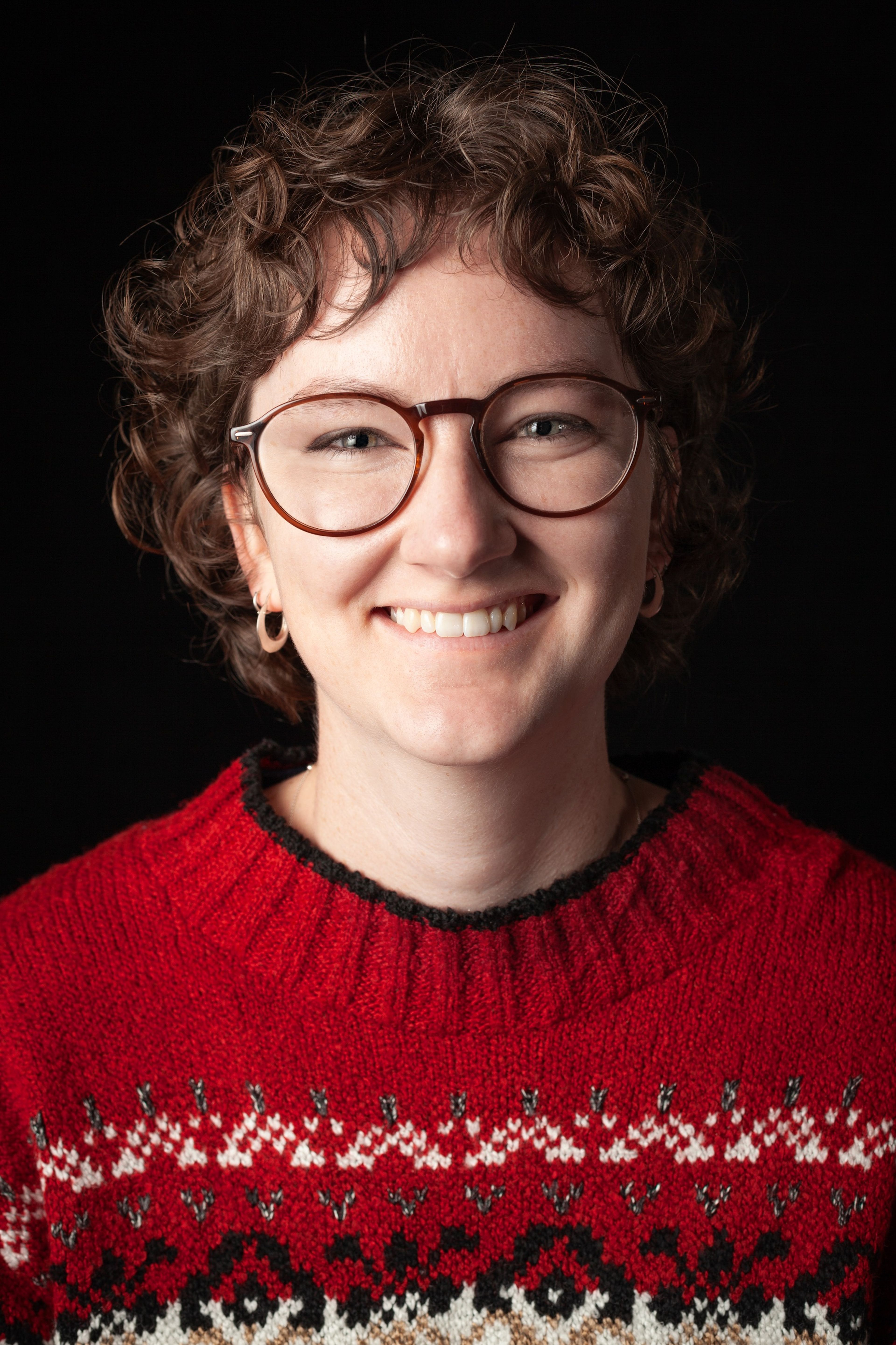 Headshot of a smiling young woman with curly brown hair, wearing glasses and a red patterned jumper, against a black background.