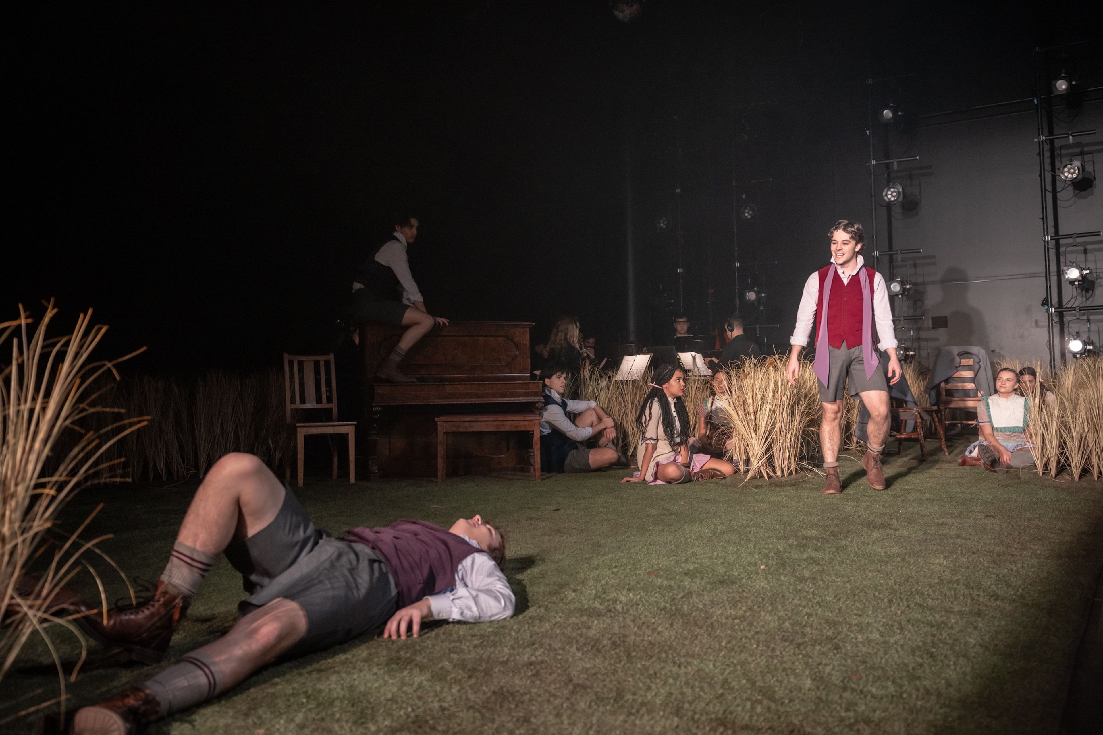 Theatre production photograph showing actors in school uniforms spread across a set designed with grass and tall grasses, evoking a naturalistic scene, with one actor standing and others sitting or lying down.