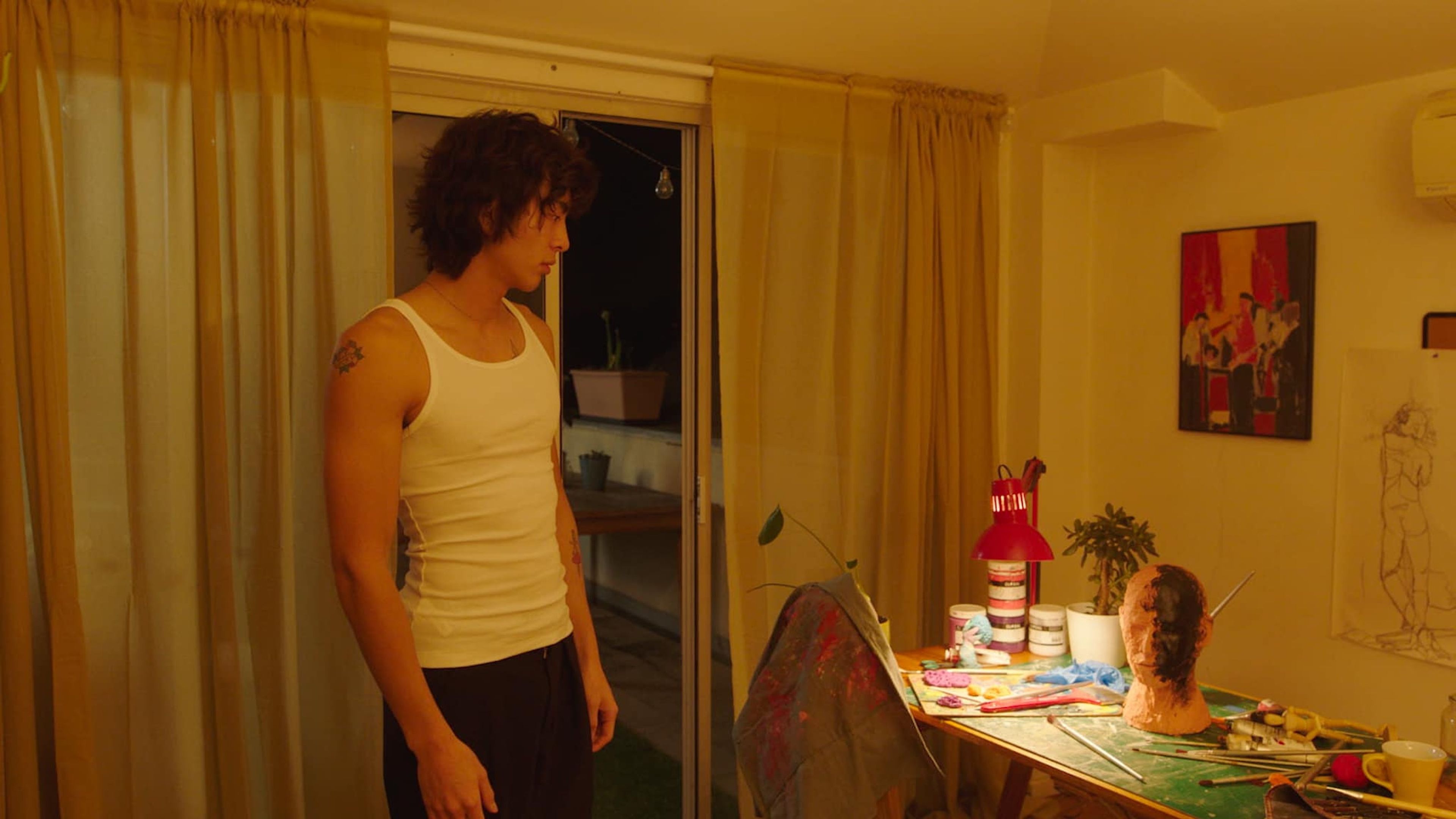 Film still showing a man in a white tank top looking towards an art workspace, with paint, tools, and a mannequin head on a table in a warm-lit room.