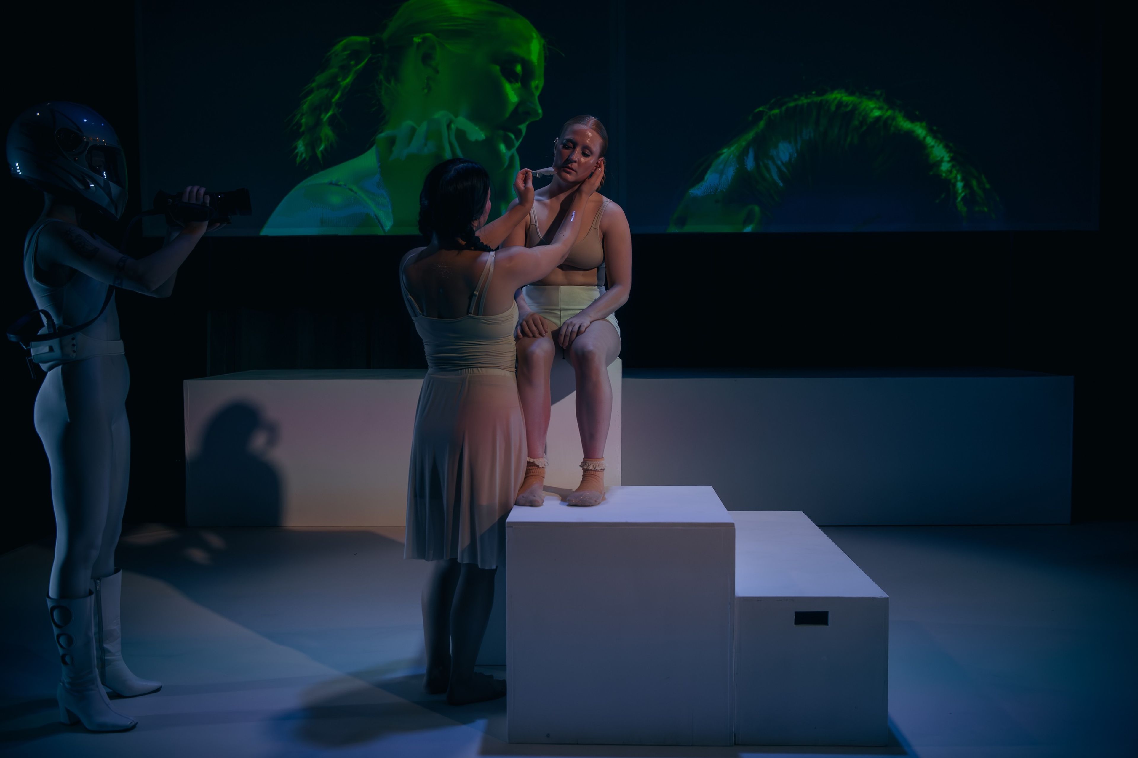 Theatre production photograph of two performers on a white platform, with one applying makeup to the other, and a projected close-up of the actor's face in green lighting behind them.