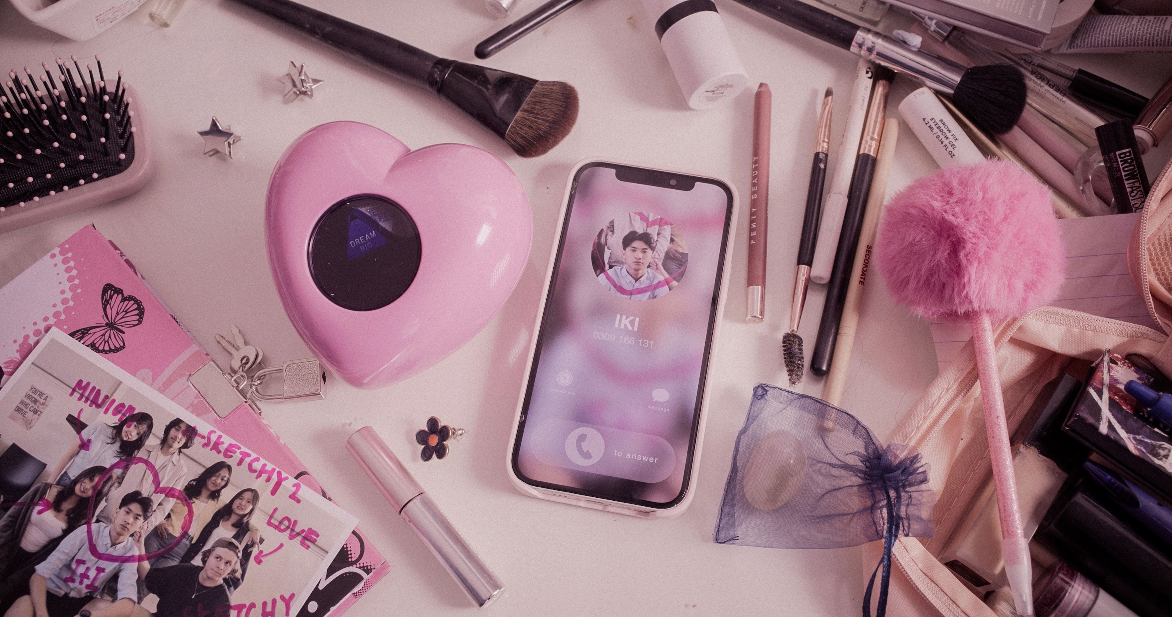 Prop styling photograph of a teenager's desk featuring pink makeup brushes, a heart-shaped speaker, phone displaying a social media profile, and K-pop fan memorabilia arranged on a pale surface