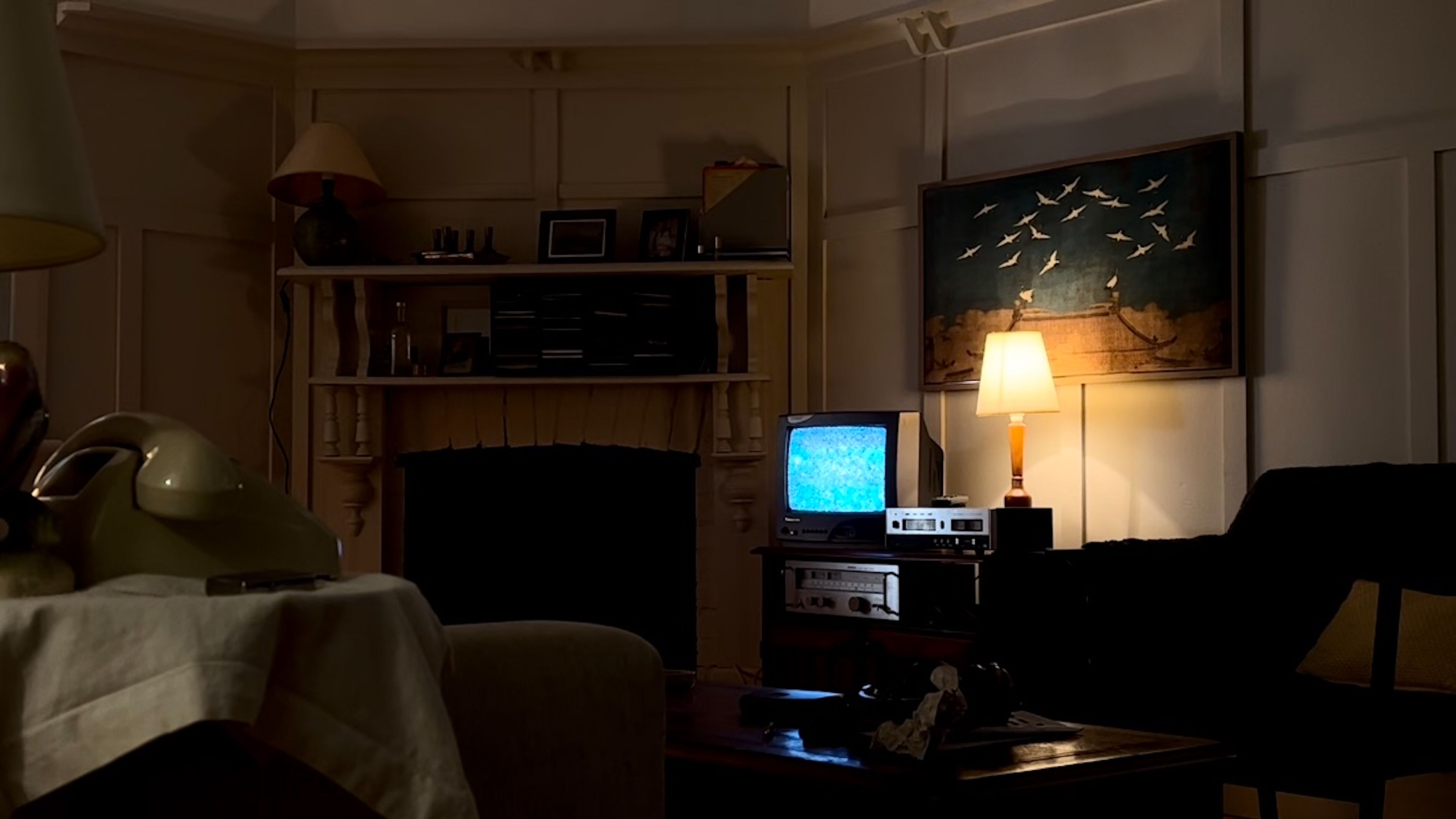 Set design photograph of a darkened 1980s living room interior featuring a vintage TV with blue screen, wooden mantelpiece, table lamp, and wall art depicting birds in flight