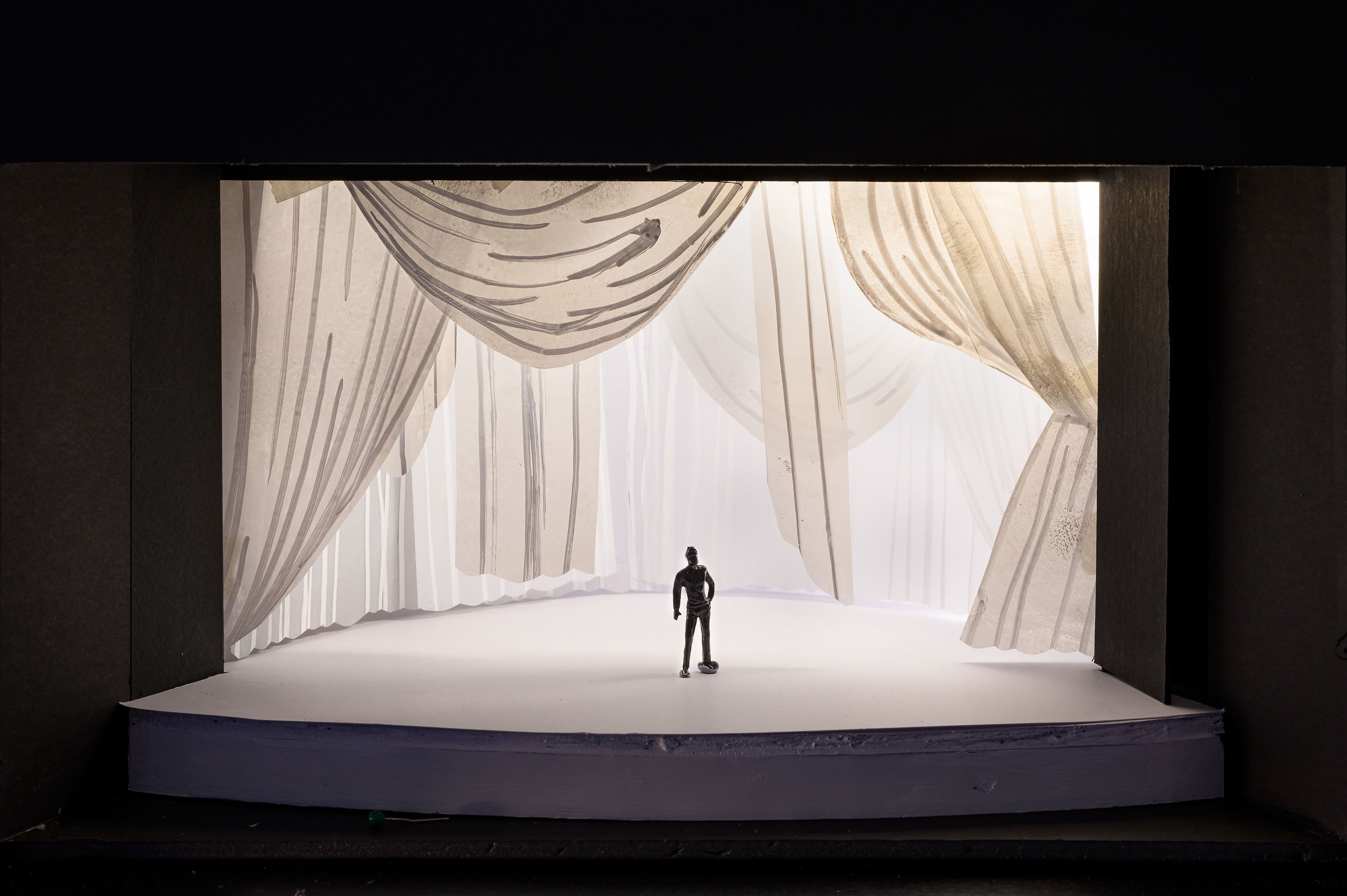 Stage model featuring soft, flowing drapery in neutral tones that cascades around the backdrop, creating a serene and ethereal ambiance. A lone human figurine stands on a clean, white platform