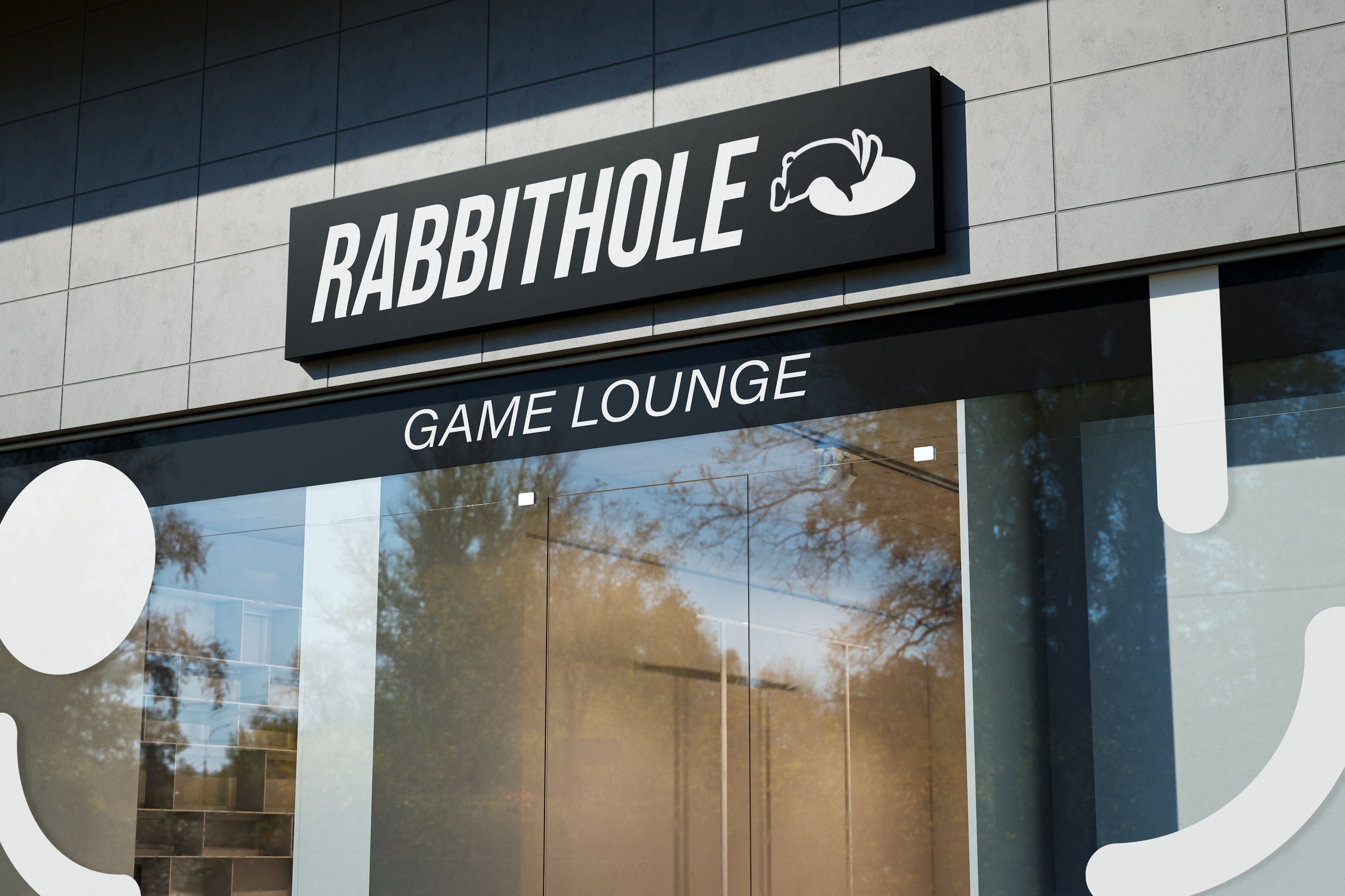 Exterior signage of 'Rabbithole Game Lounge' featuring a modern, black sign with a minimalist rabbit logo and bold, white typography. The reflective glass storefront shows faint reflections of trees and the surrounding environment.