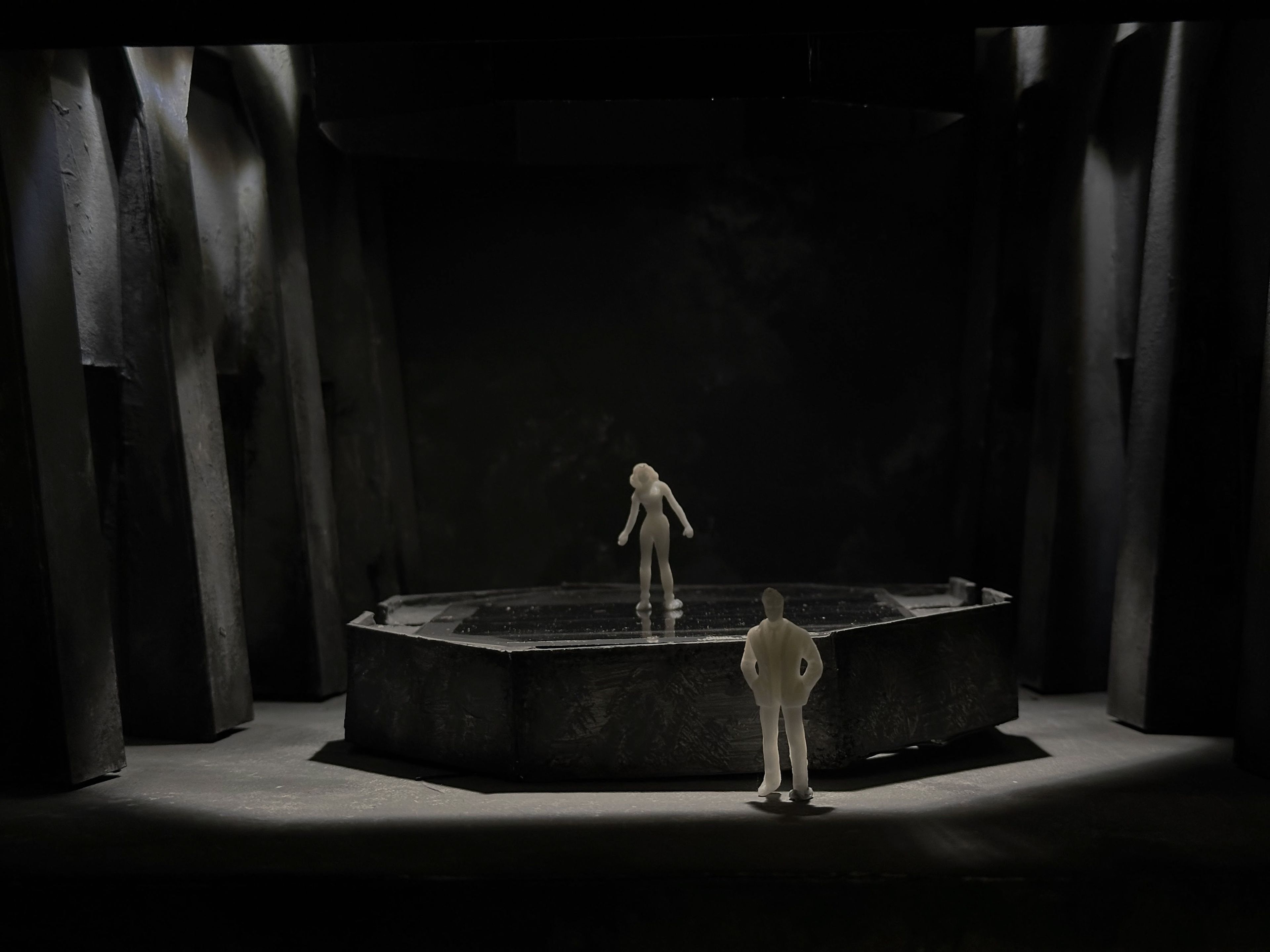 Set model with two figures facing each other on a hexagonal platform surrounded by columns, lit dramatically from above.