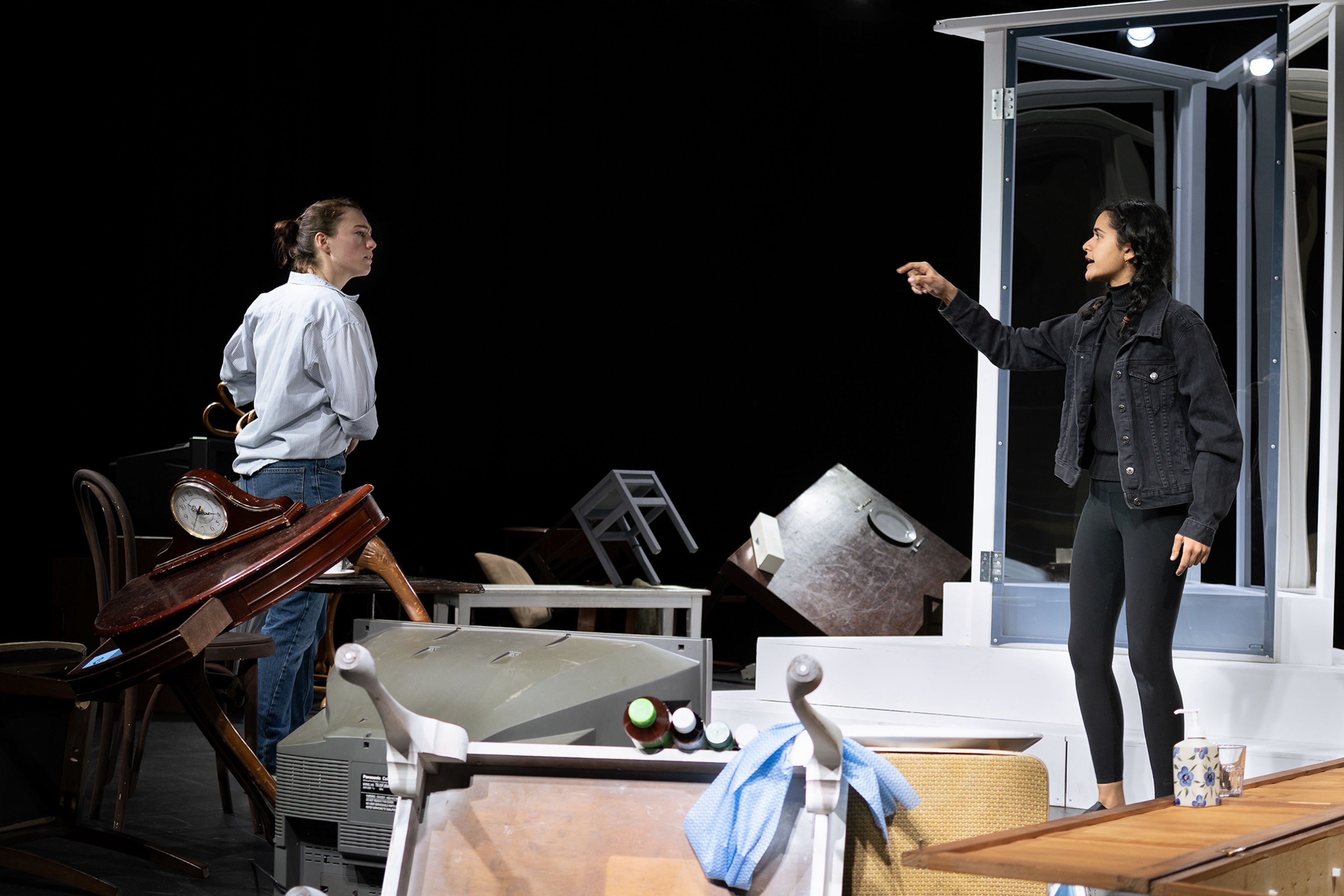 Theatre production photograph showing two performers, one in a white shirt and jeans near piled wooden furniture, the other in black clothing standing in a doorframe, on a stark stage.