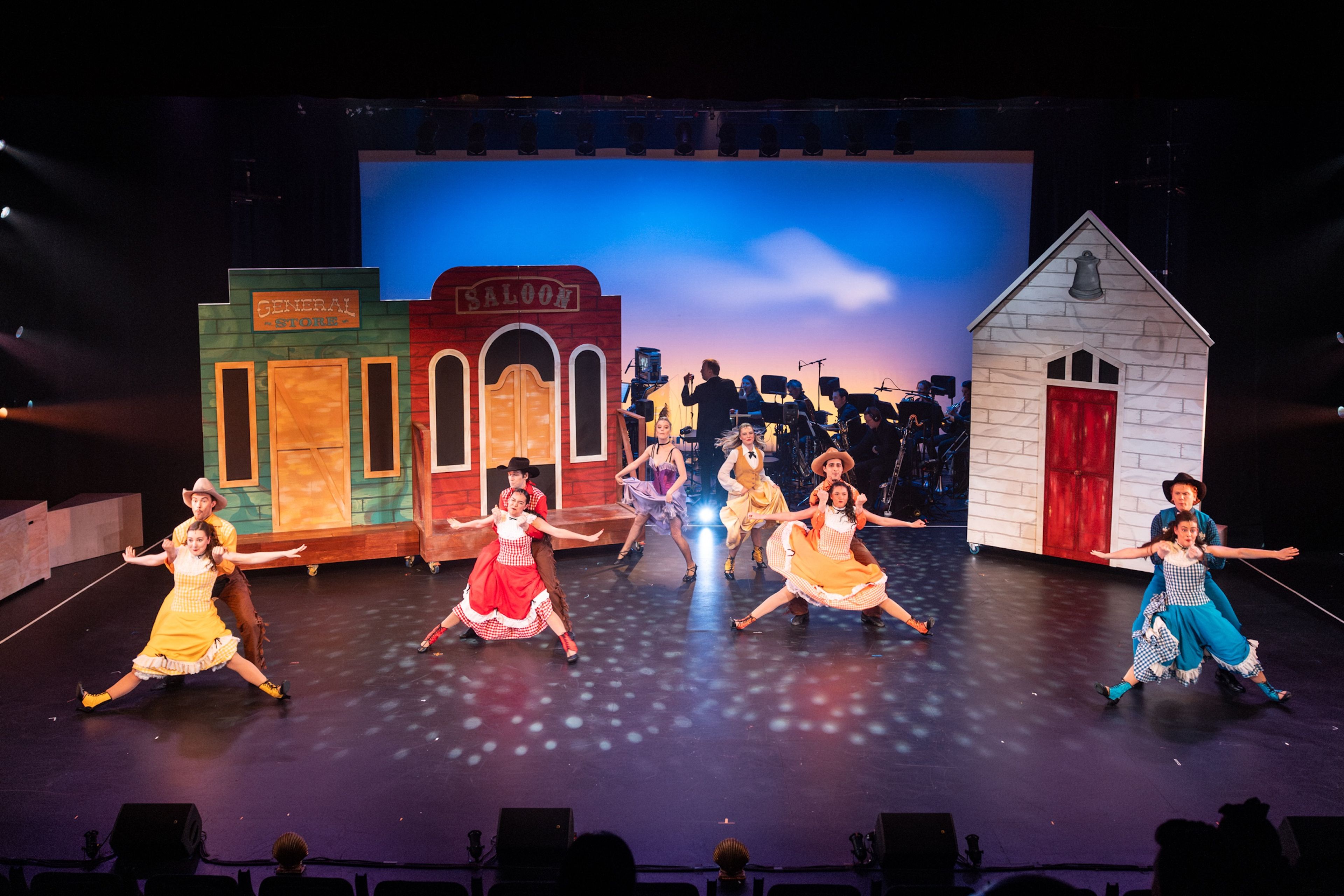 Theatre production photograph with performers in colourful western-themed costumes, dancing in pairs against a set with a saloon and general store facade, under bright stage lighting with a blue sky backdrop.