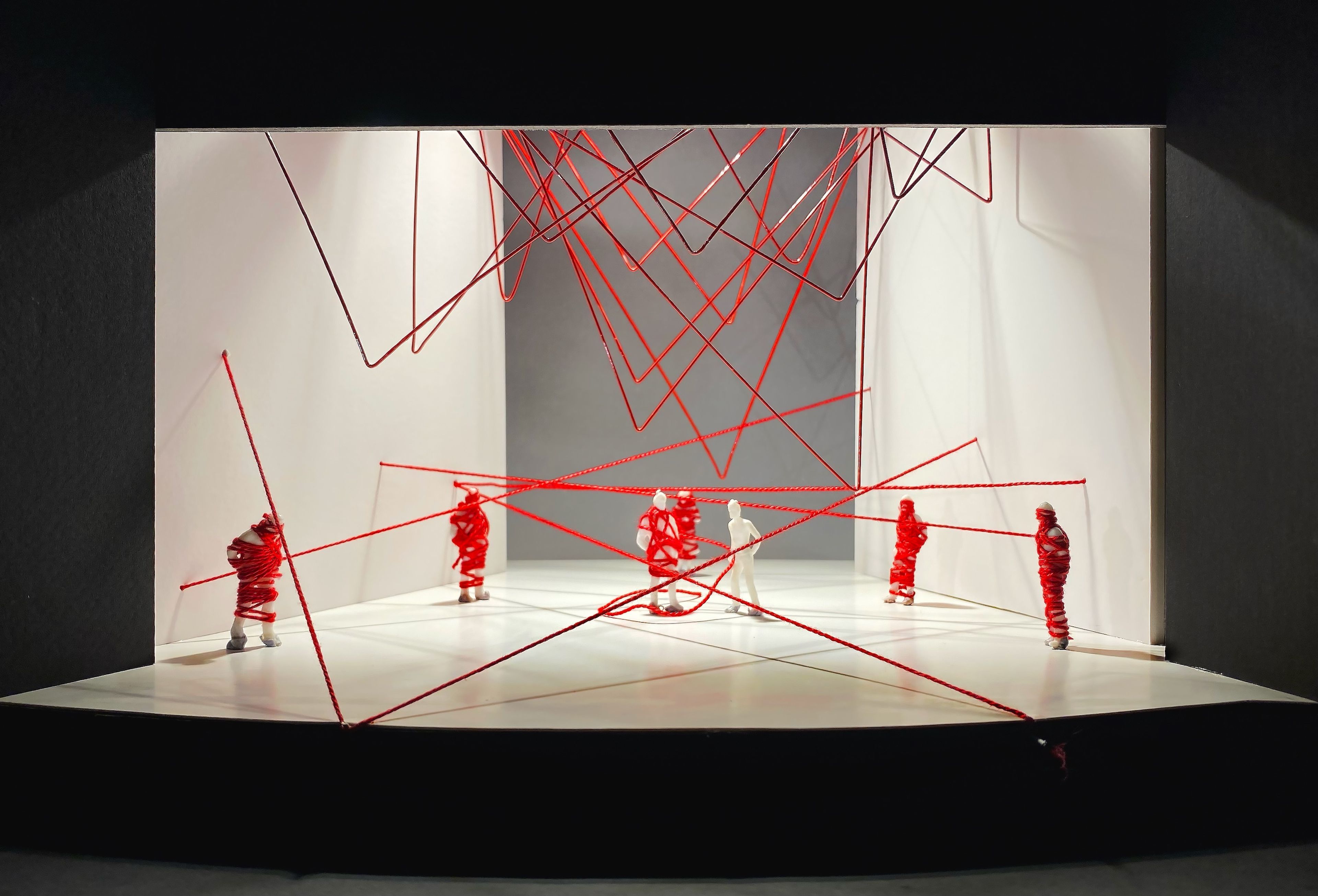 A set model with white walls and figures wrapped in red thread. The set features intersecting red strings suspended from above, creating a web-like pattern across the space. Small human figures are positioned in various parts of the stage, wrapped tightly with the red thread.