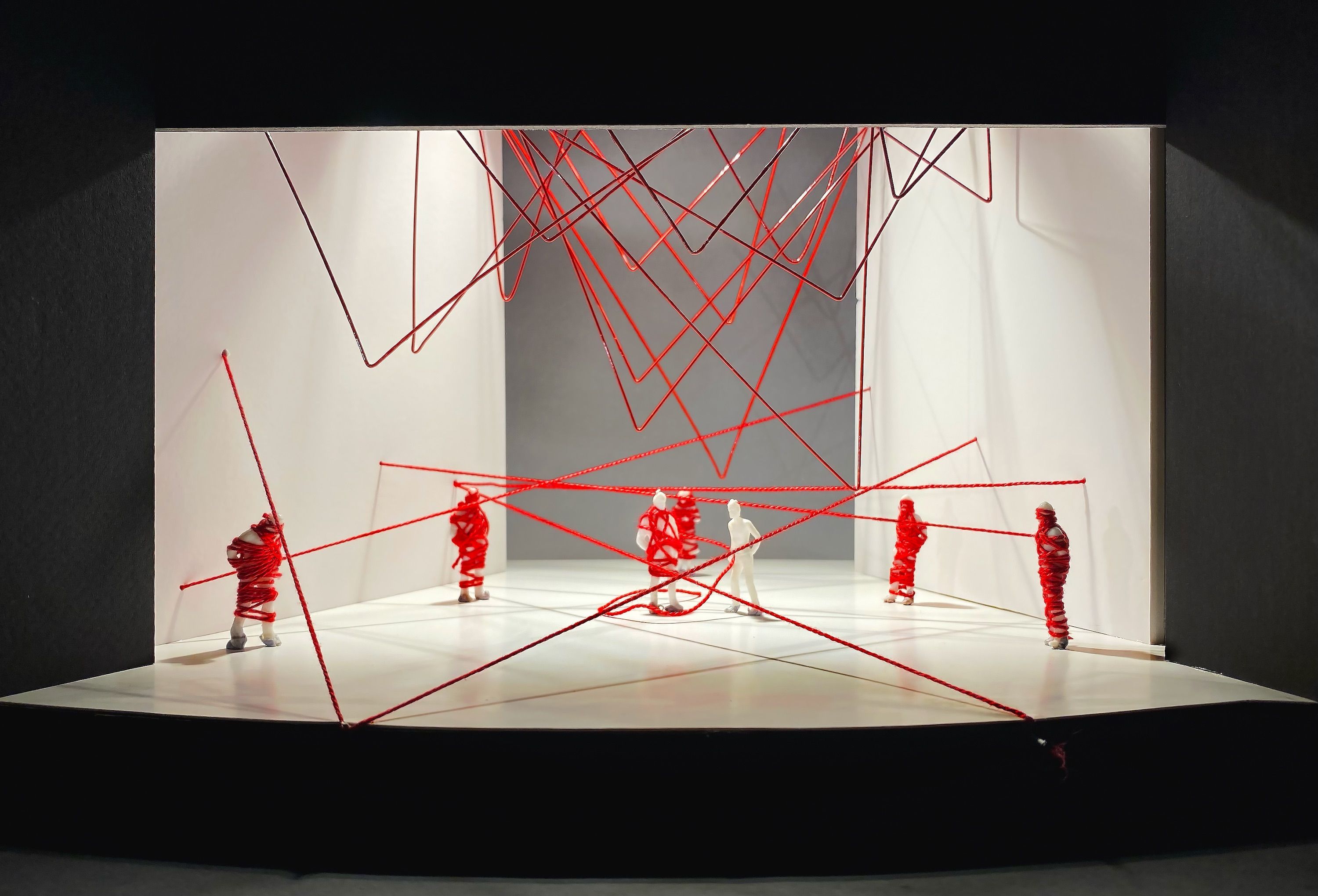 A set model with white walls and figures wrapped in red thread. The set features intersecting red strings suspended from above, creating a web-like pattern across the space. Small human figures are positioned in various parts of the stage, wrapped tightly with the red thread.