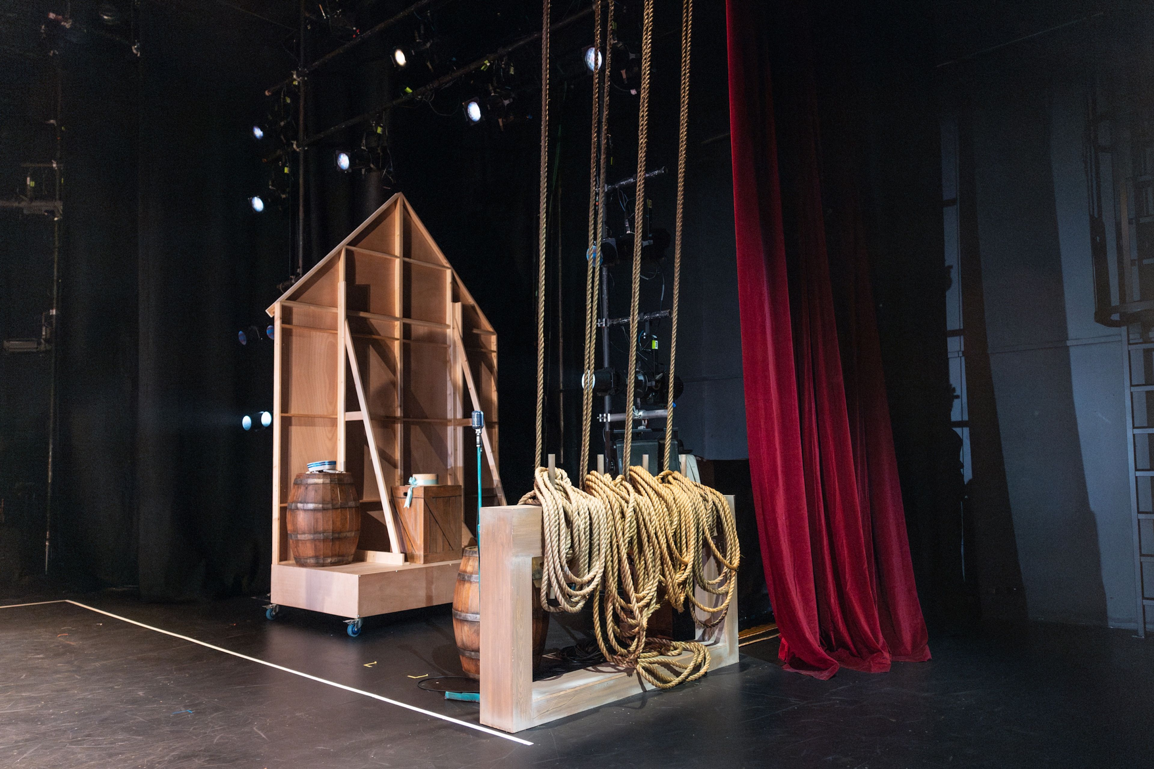 Backstage setup featuring a stage prop resembling a wooden shack and a large collection of coiled ropes hanging near a red curtain, creating an industrial atmosphere.