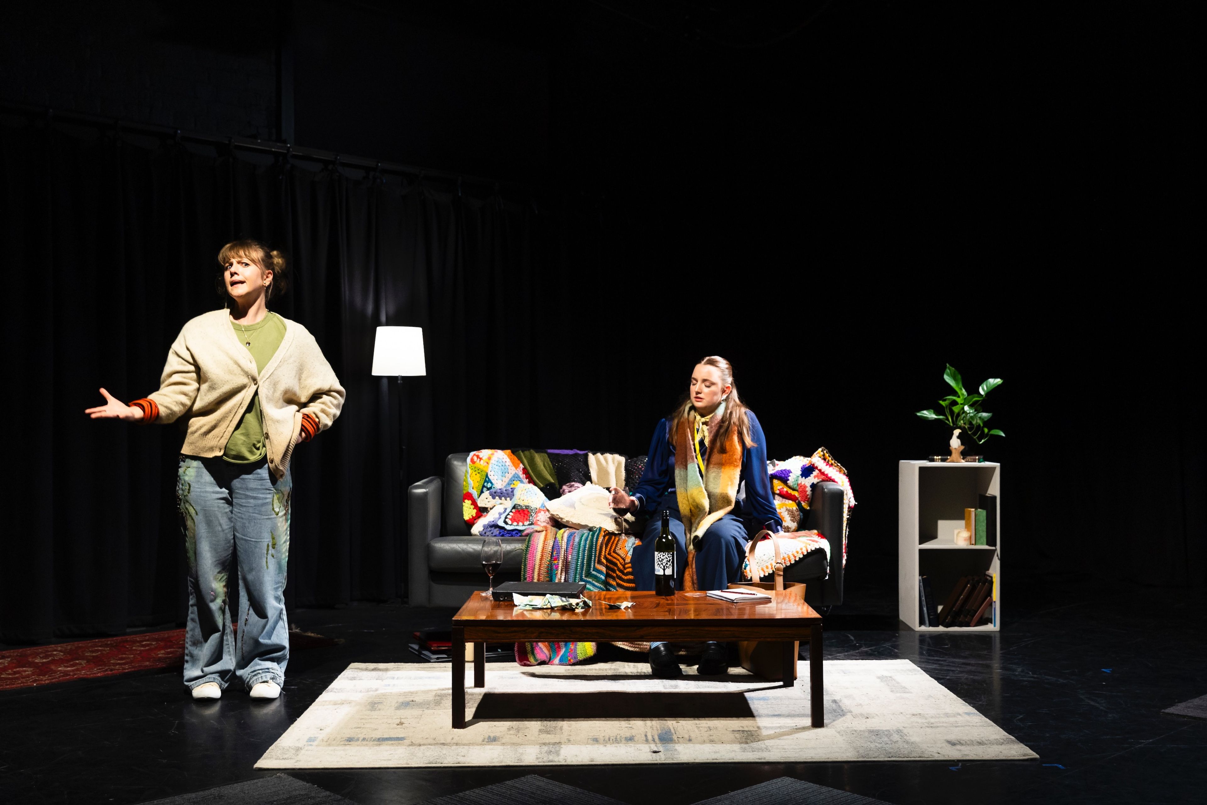 Two actors on stage in a living room setup, one standing and gesturing while the other sits on a couch covered in a colourful knitted blanket, evoking a cosy domestic atmosphere.