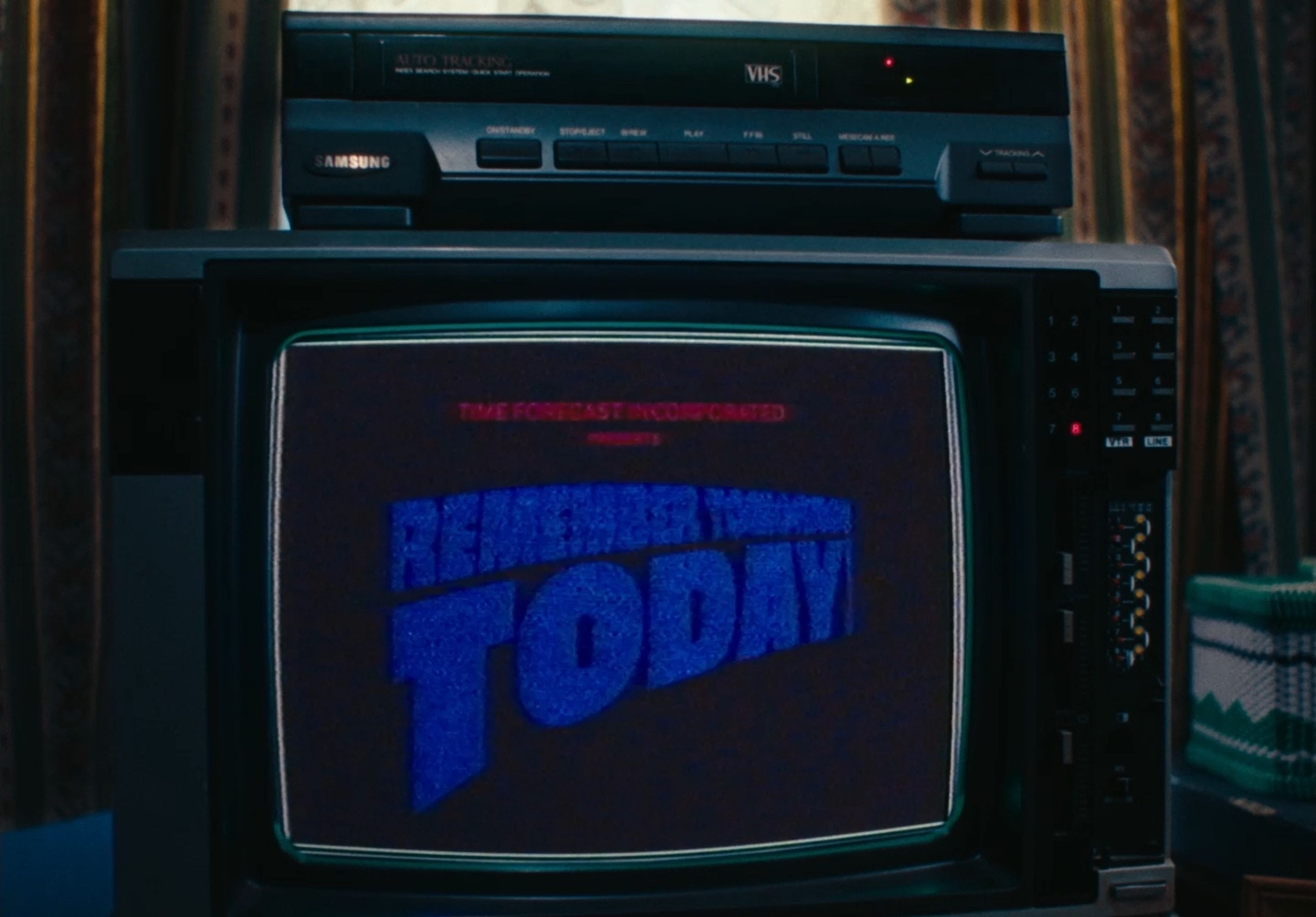 Film still showing a Samsung VHS player and TV screen displaying the 'Remember Tomorrow Today' program logo in bright blue text against a dark background.